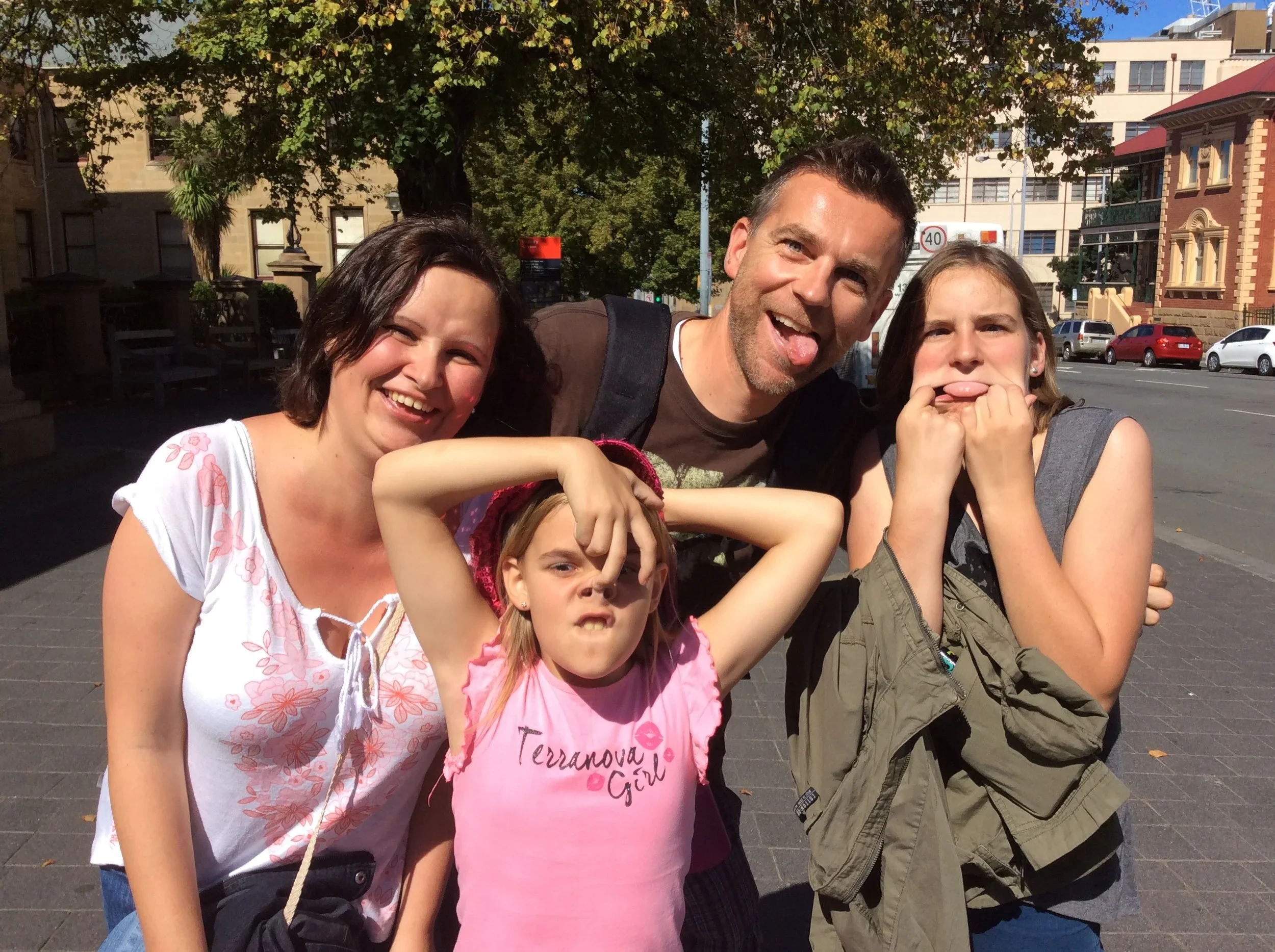 A group of five people, two adults and three children, posing for a photo outdoors on a sunny day, making funny faces and gestures.