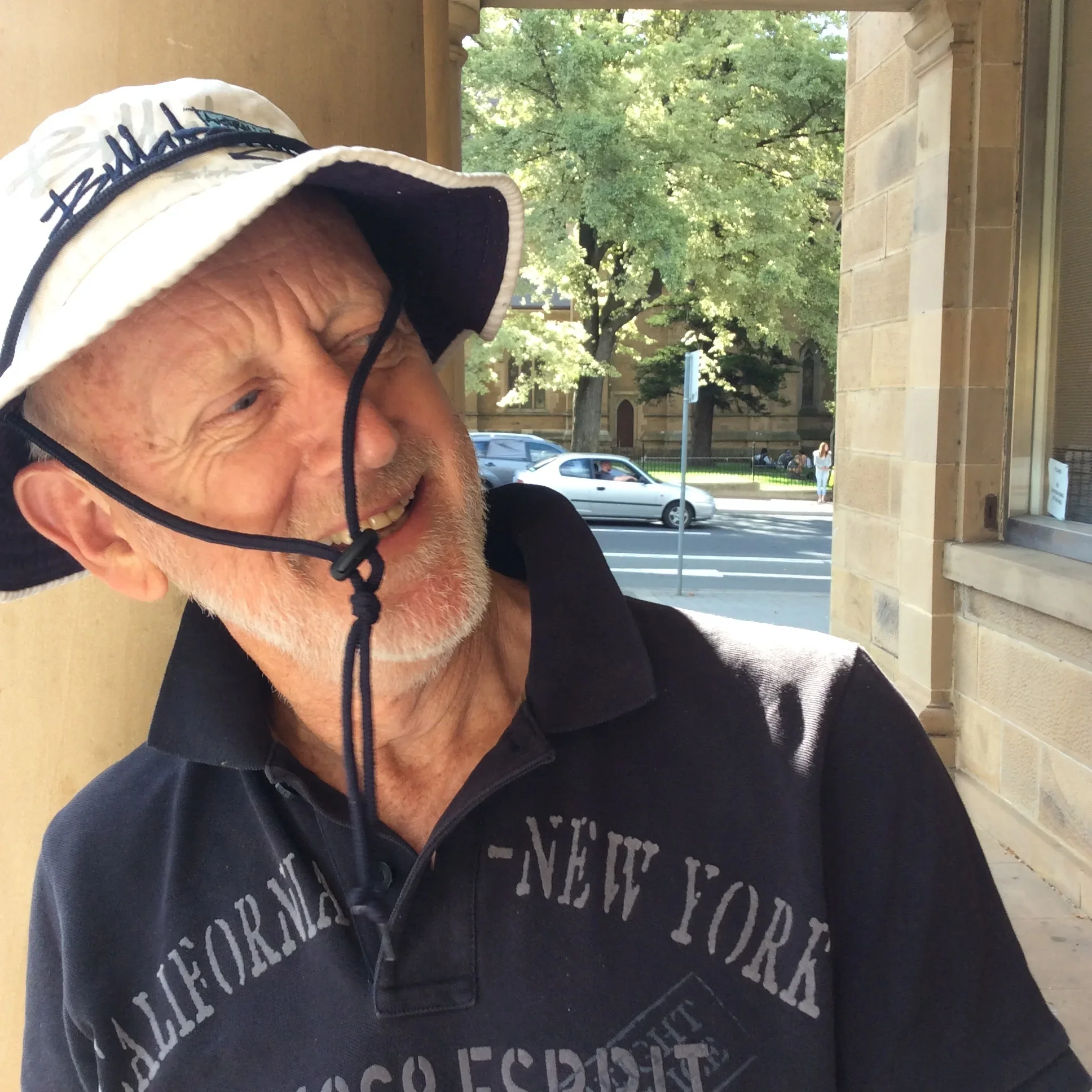 An elderly man wearing a wide-brimmed hat and a black polo shirt with 'California - New York - Los Angeles' printed on it, standing outside a building with a street scene and trees in the background.