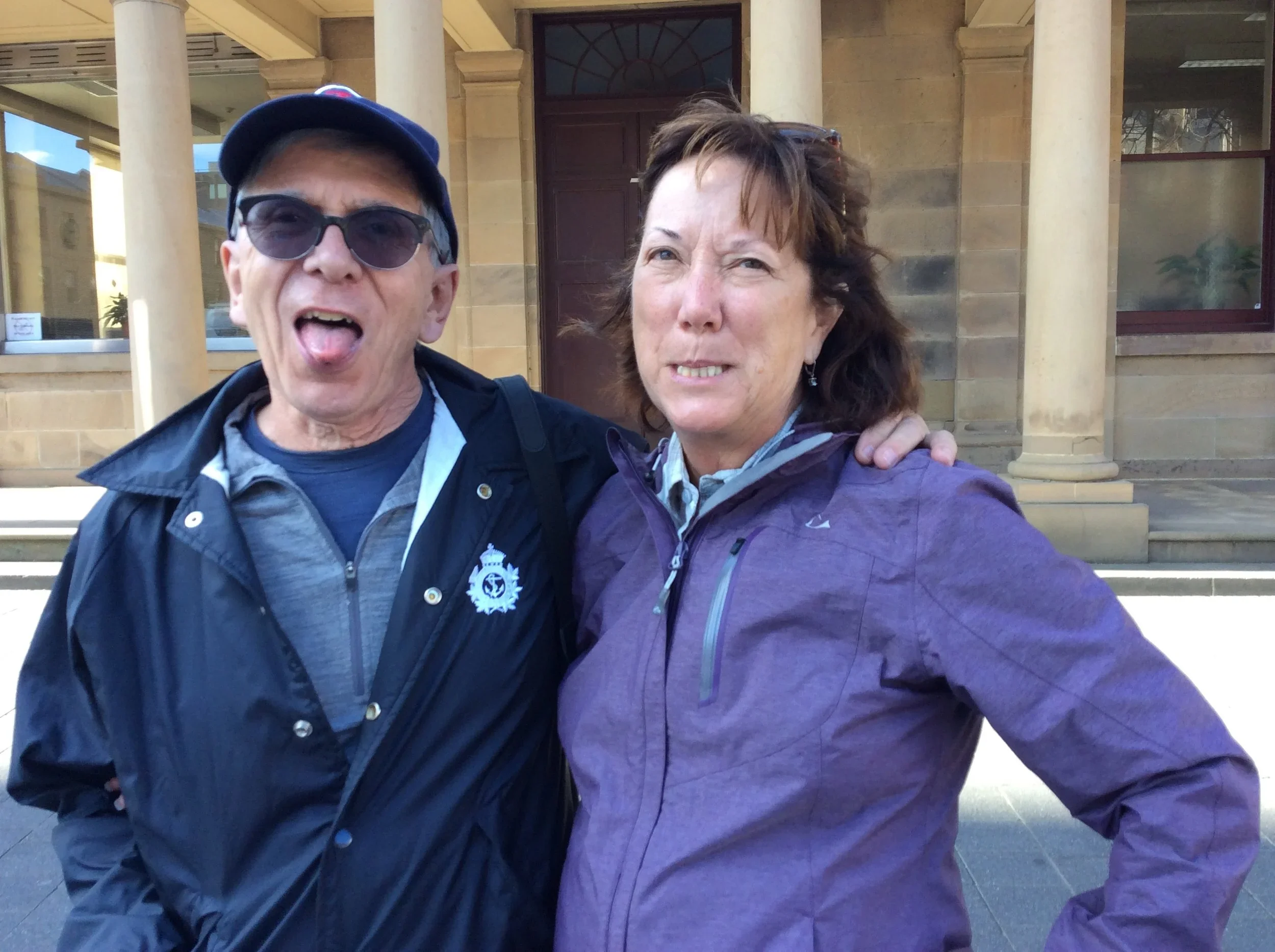 A man and woman posing together outside a building, with the man sticking out his tongue and the woman looking serious. The man is wearing sunglasses, a dark jacket, and a cap, while the woman is wearing a purple jacket and earrings.