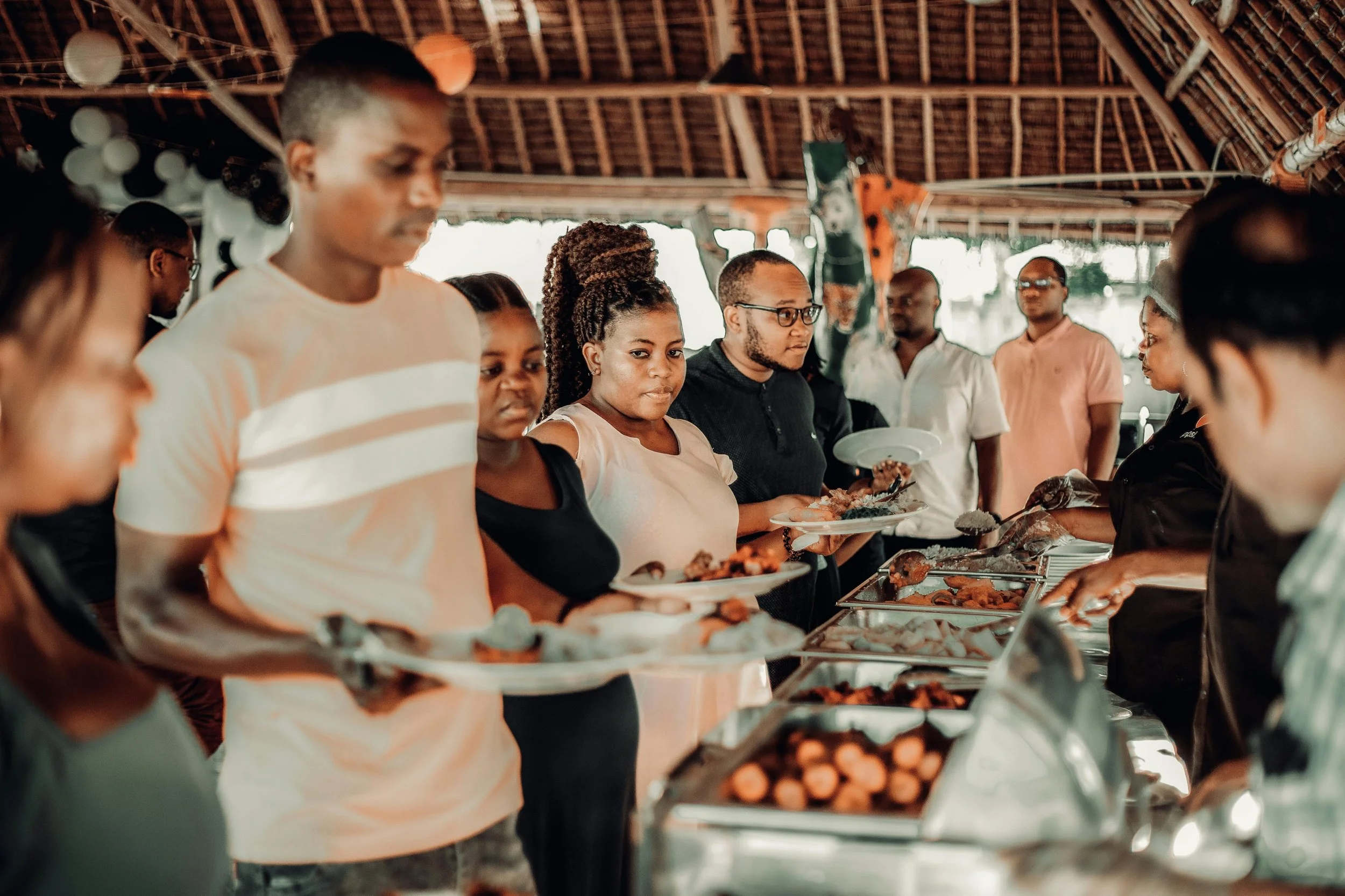 A community outdoor picnic and barbecue event with many people, including children and adults, gathered around long tables with food, in a park near a basketball court and playground, under a white canopy tent with a banner that reads 'The Kin Connection 2026'.