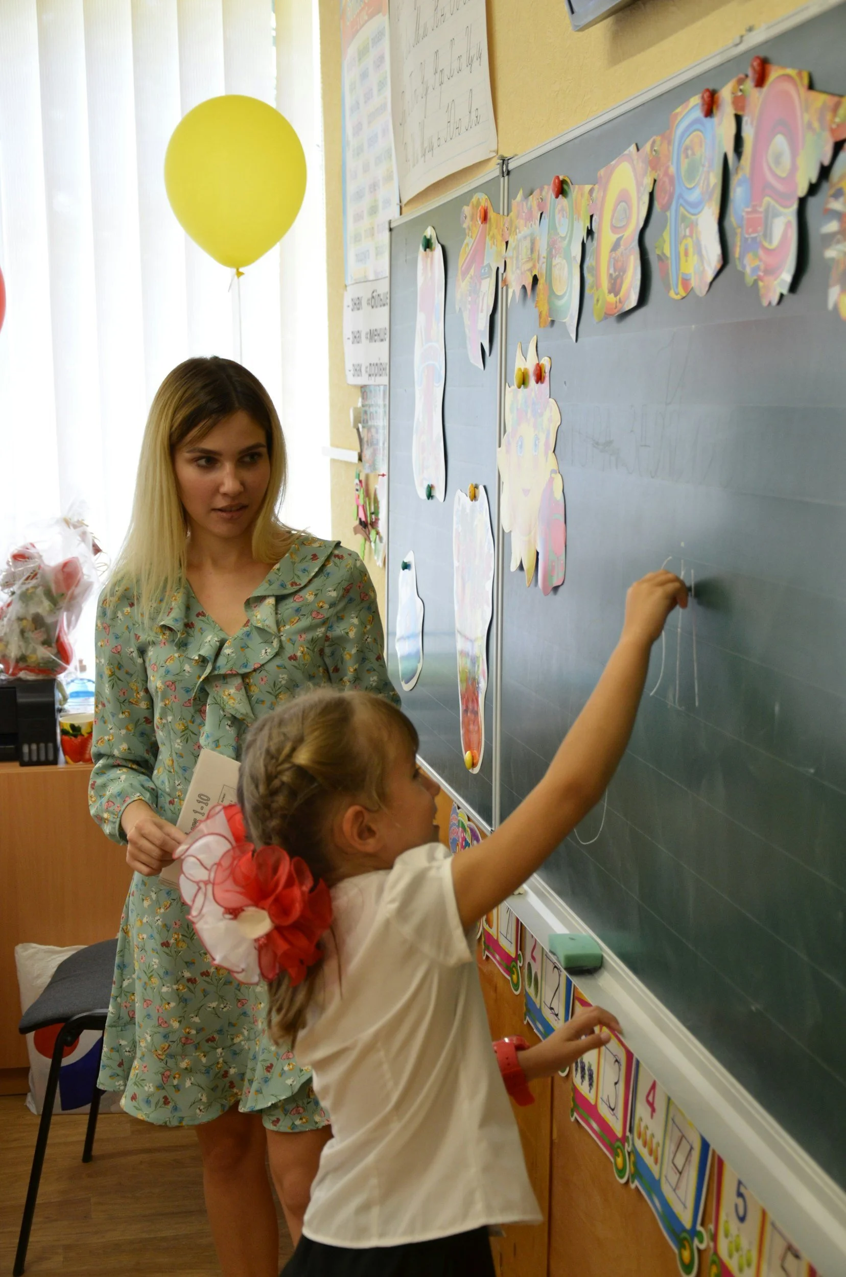 A classroom scene with a teacher observing two young girls drawing on a chalkboard. One girl is wearing a white shirt with a red flower accessory in her hair, and the other girl has braided hair and a red bow. A yellow balloon is floating near the window, and colorful alphabet and number posters are on the wall.