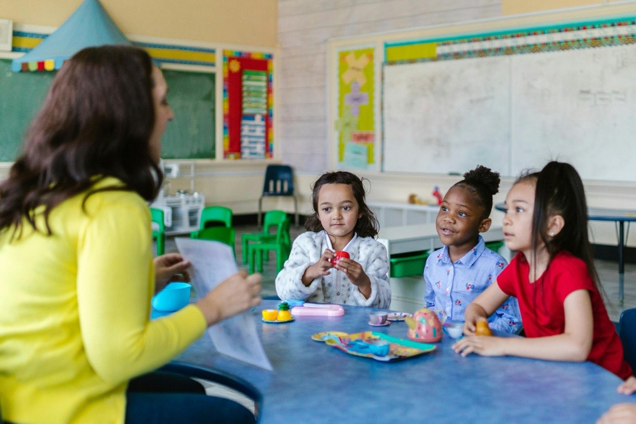 A teacher sitting at a table with three young girls in a classroom, engaging in a teaching activity with toys and play items on the table.