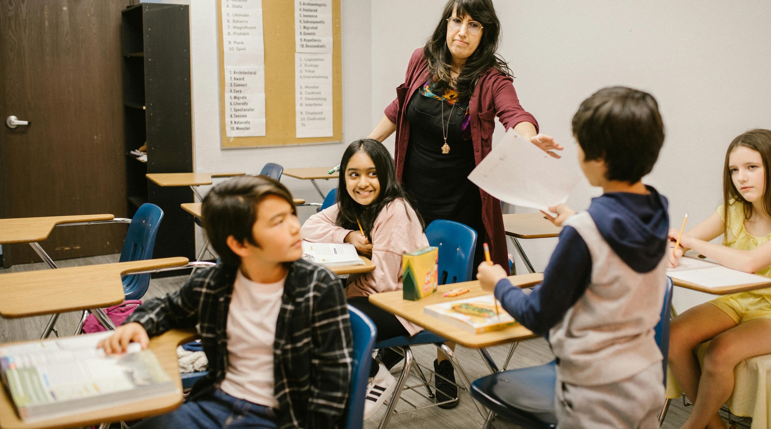 A teacher and a group of young students in a classroom, with some sitting at desks and others standing, engaged in a learning activity.