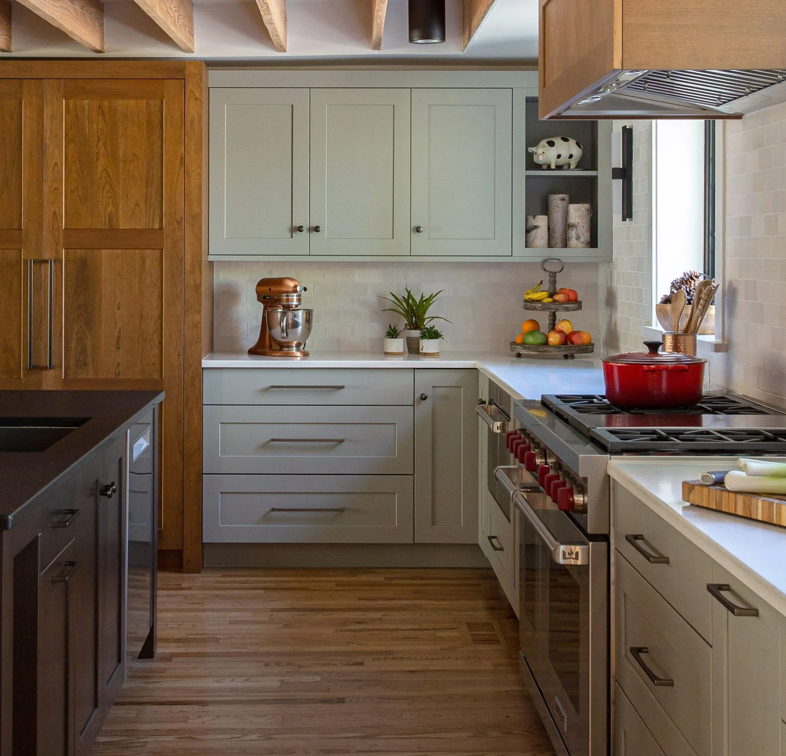 Modern farmhouse open plan kitchen with beamed ceilings, wood floors, mix of cherry and green cabinets and large island. Sub-Zero paneled refrigeration is hidden and the Wolf 48” range is featured.