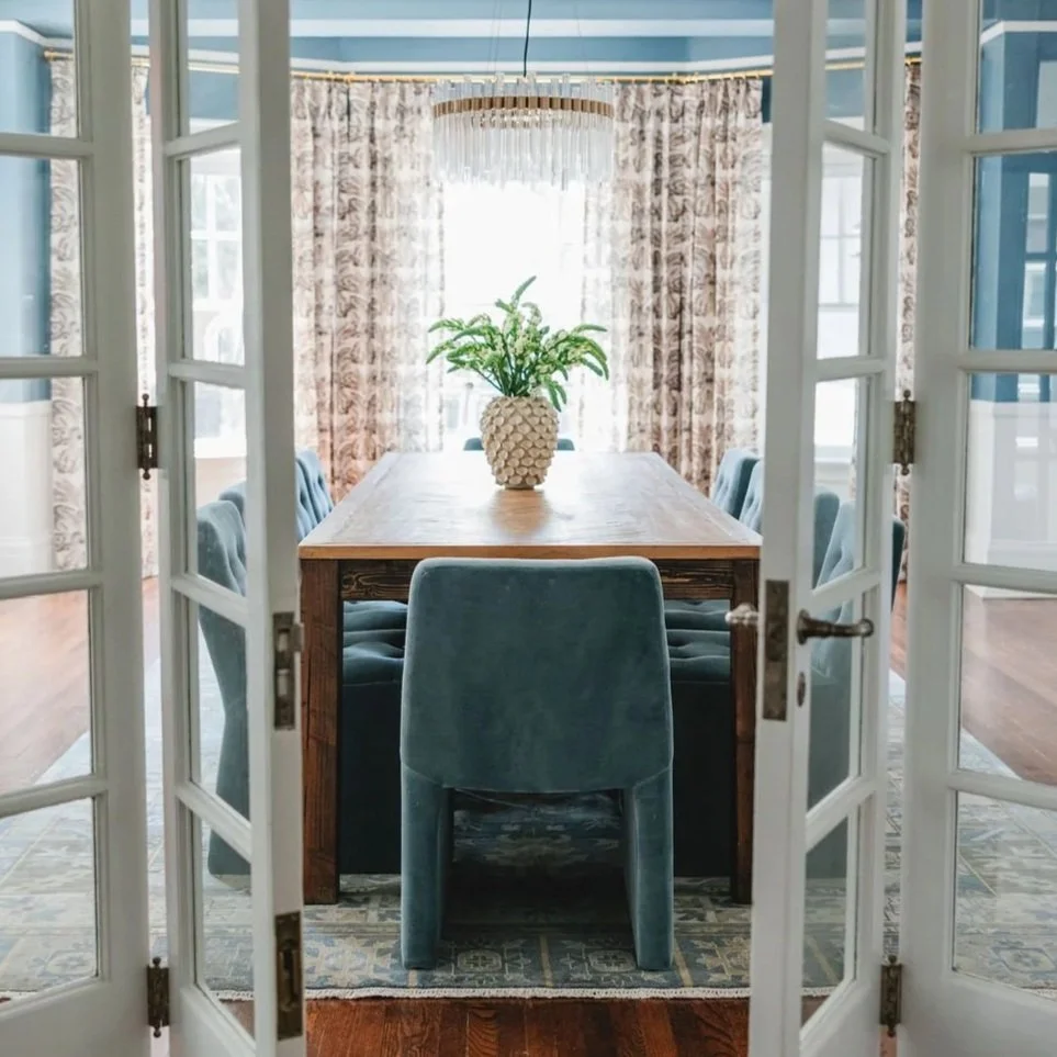 Detroit historic home dining room with a wooden table, six blue upholstered chairs, a plant in a textured white vase, and patterned curtains, viewed through white-framed glass double doors.