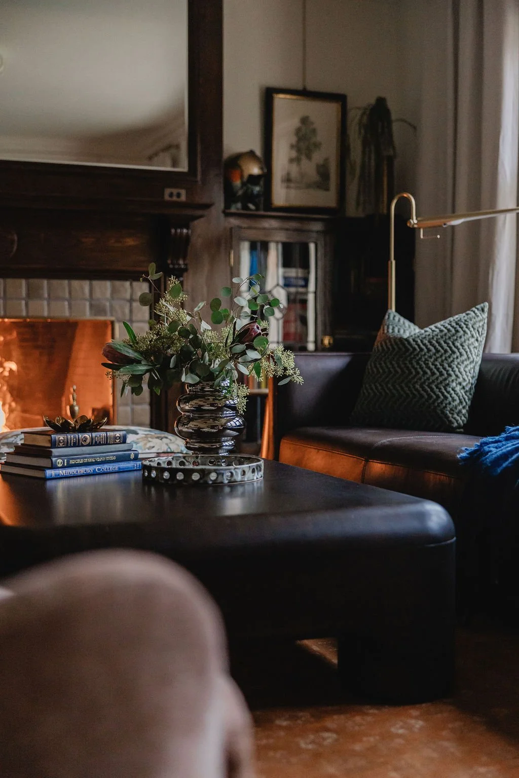 A cozy Detroit-area historic living room featuring a black leather sofa with patterned cushions, a wooden coffee table with a floral arrangement, stacked books, and a fireplace with a mirror above it. The room has warm and unique interior design.