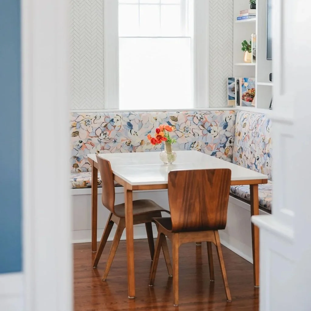 A cozy Detroit breakfast nook with a white marble table, surrounded by wooden chairs, and a built-in floral-patterned banquette cushion with pastel colors. Historic home interior design