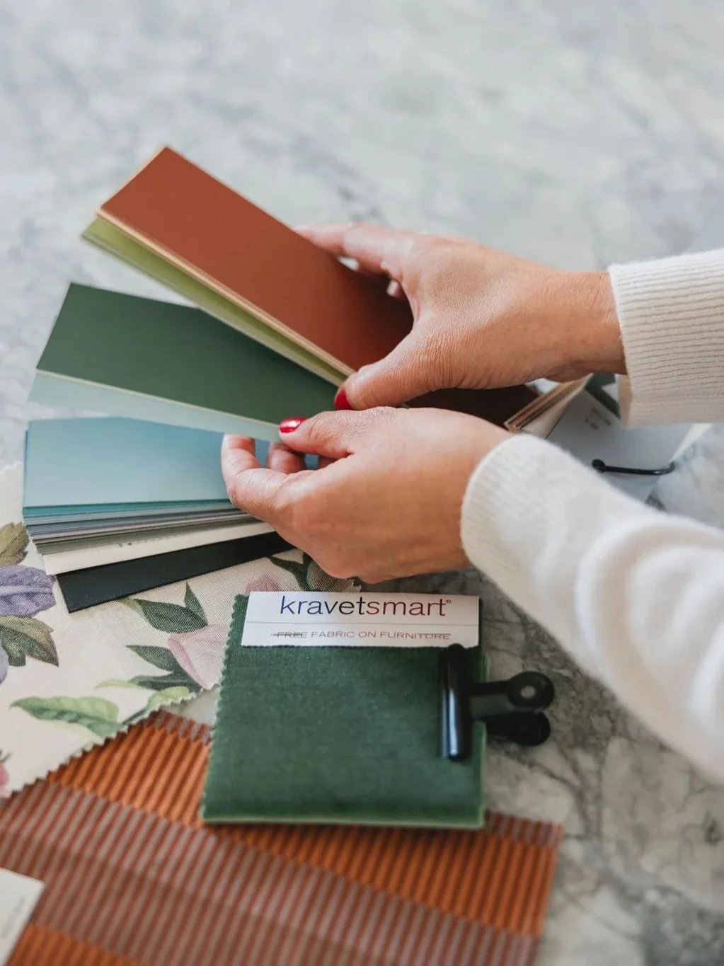 Person holding color swatches in shades of brown, green, and blue, with fabric and paint samples on a table.