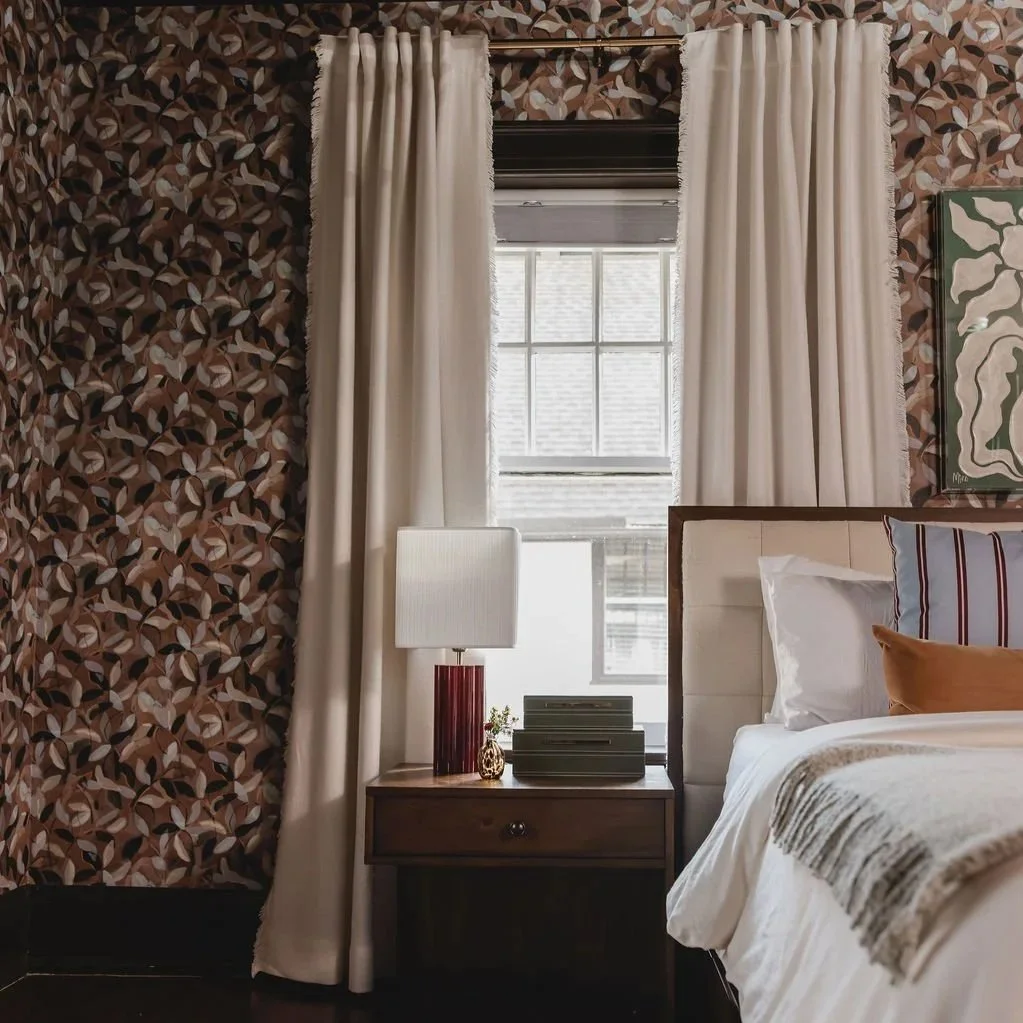 A Detroit bedroom corner with a window, white curtains, a bedside table with a lamp, and a bed with pillows and a blanket. Historic home interior design.