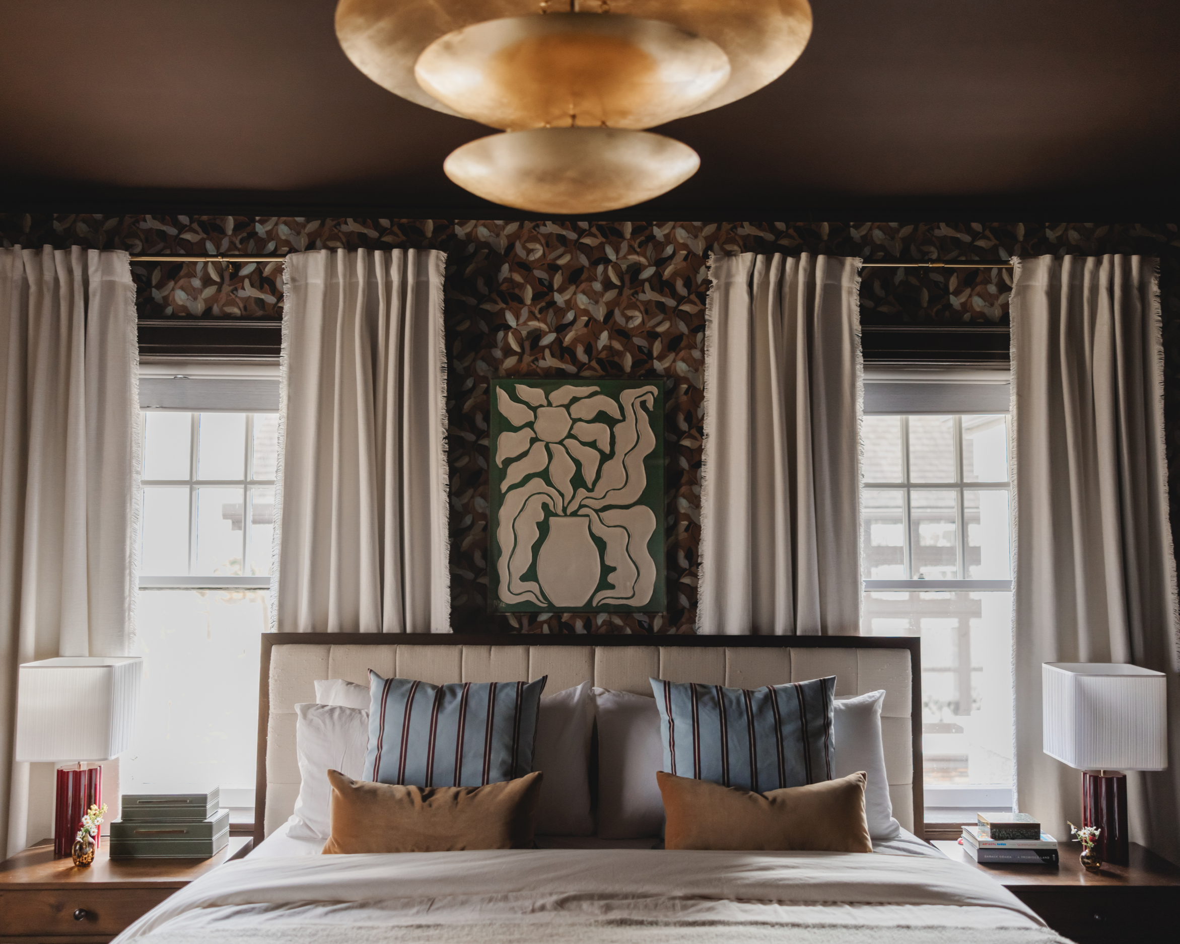 A moody Detroit bedroom with a bed against a floral wallpapered wall, framed by two large windows with white curtains, featuring two bedside tables with lamps, books, and small decorative items.