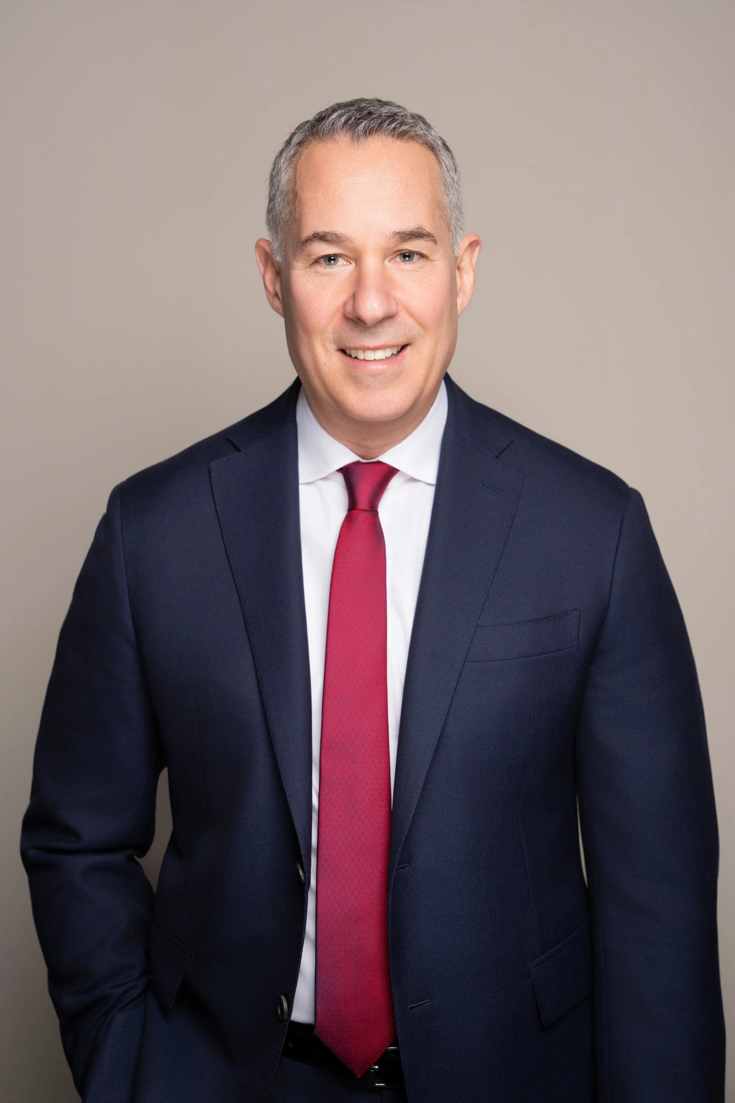 Professional portrait of Justin V. Shur, wearing a dark blue suit, white dress shirt, and red tie, standing against a neutral background.