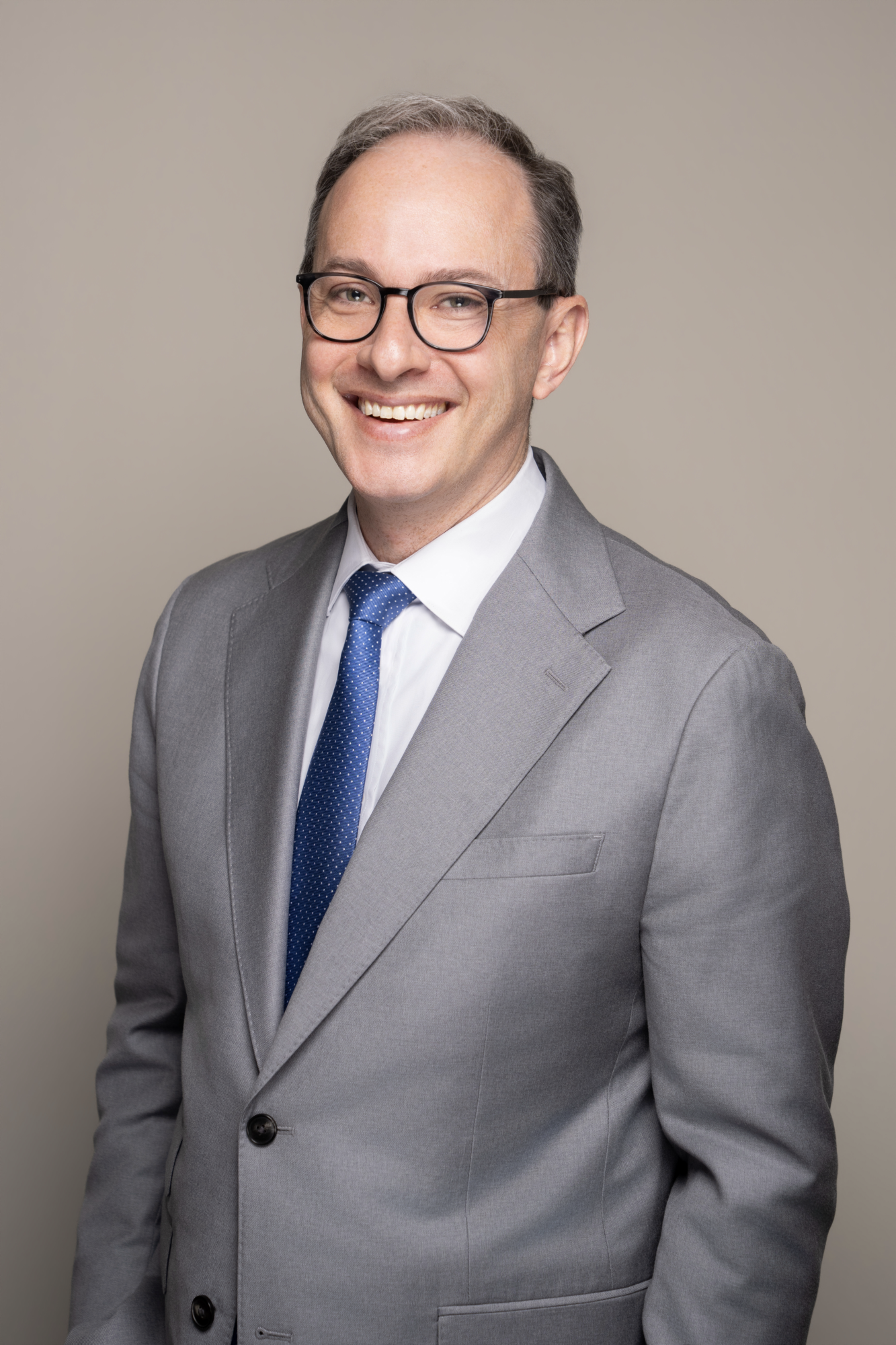 Professional portrait of Jonathan I. Kravis in a gray suit, white shirt, and blue tie, smiling and wearing glasses, against a plain beige background.