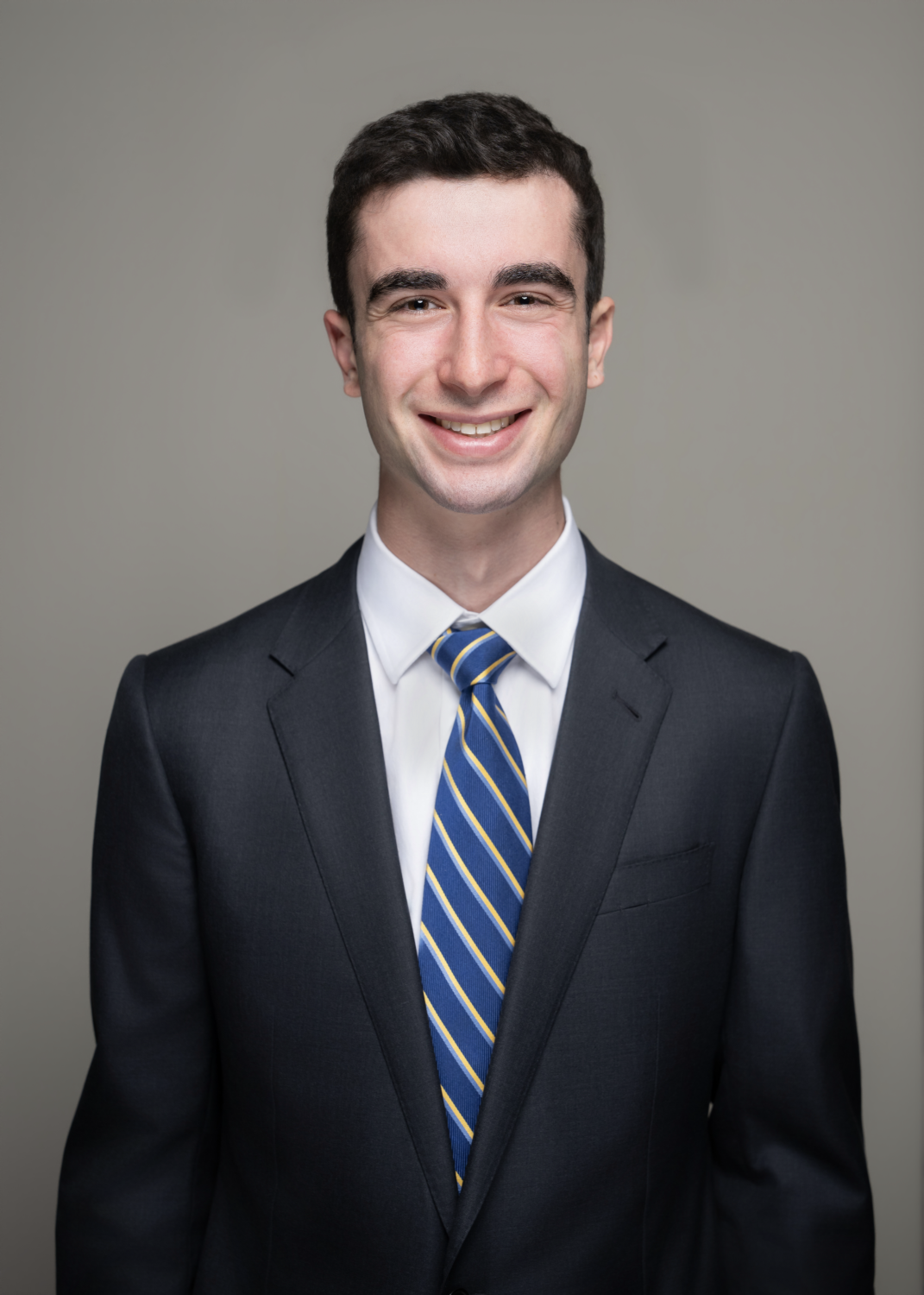 A young man with dark hair, smiling, dressed in a dark suit, white shirt, and a blue striped tie, standing against a plain gray background.