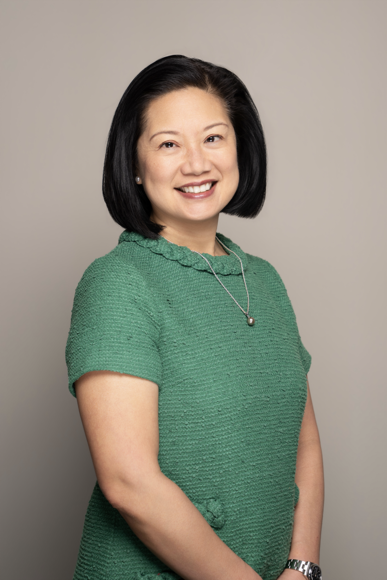 Professional portrait of Jessie K. Liu wearing a green textured dress, a silver necklace with a pearl pendant, pearl earrings, and a silver watch, standing against a neutral background.