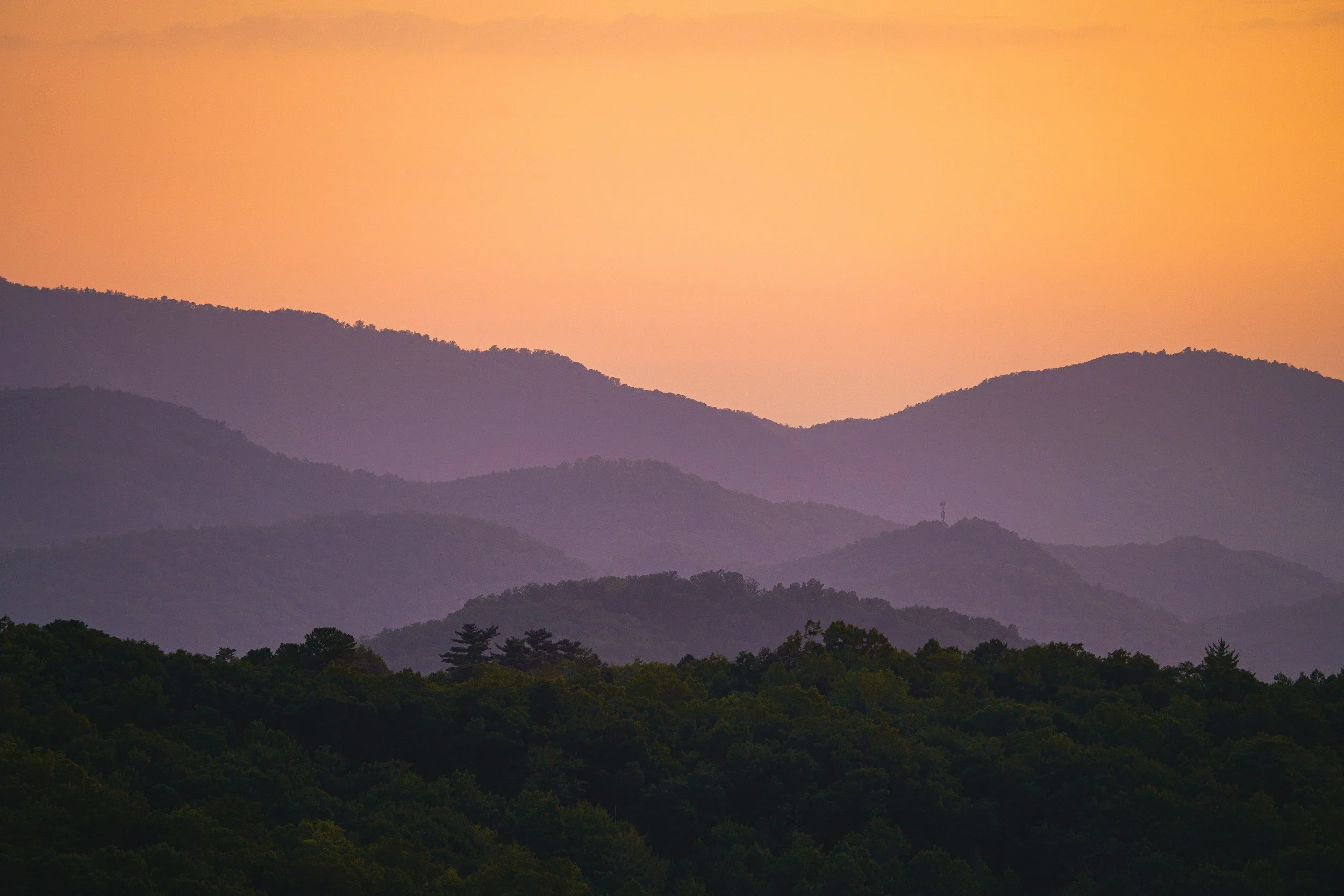 Layered Smoky Mountain silhouettes during a sunset or sunrise, with a gradient sky transitioning from orange to just a touch of pale pink. The mountains appear lavender in the mist.