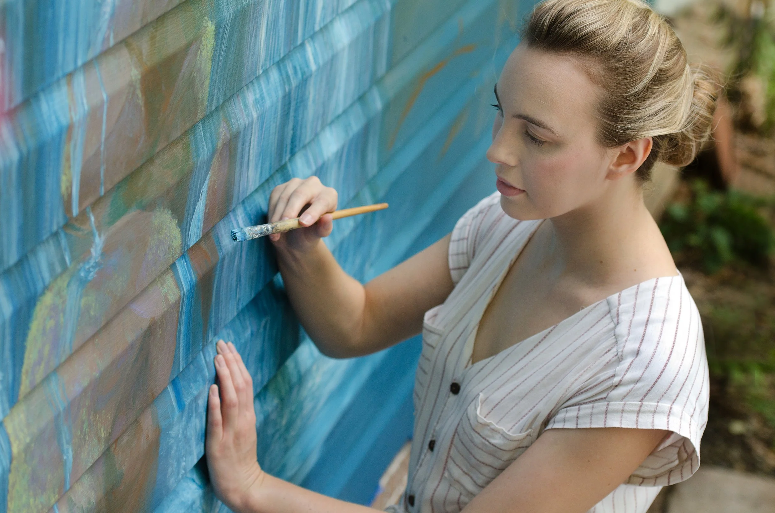 A woman painting a mural of a waterfall on a exterior wall, using a brush and blue paint.