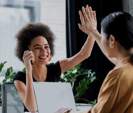 Two women smiling and high-fiving each other in an office setting. One woman is holding a phone, and the other is sitting with a laptop.