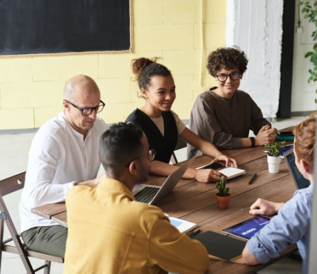 Group of five diverse young professionals sitting around a table in a meeting room with laptops and tablets.