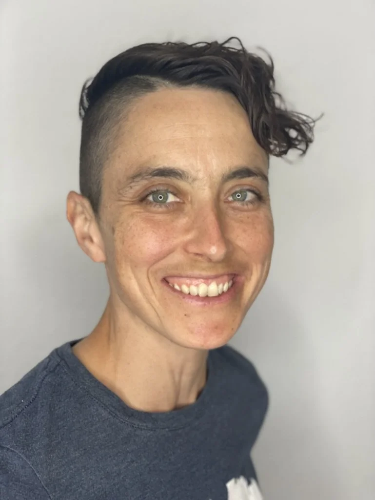 image of Elana Story smiling standing in front of light gray background wearing a grey/blue tee shirt