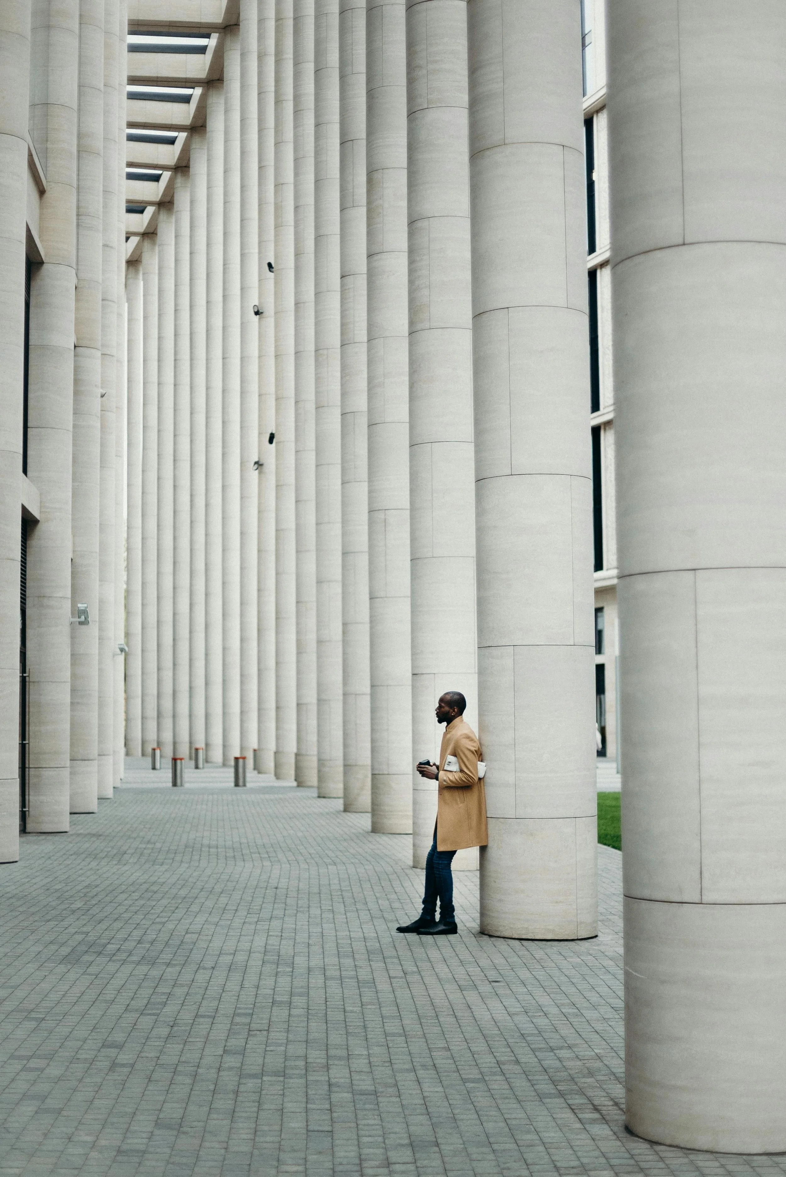 A man in a tan coat and dark jeans leaning against a large white column of a modern building with a series of similar columns extending into the distance.