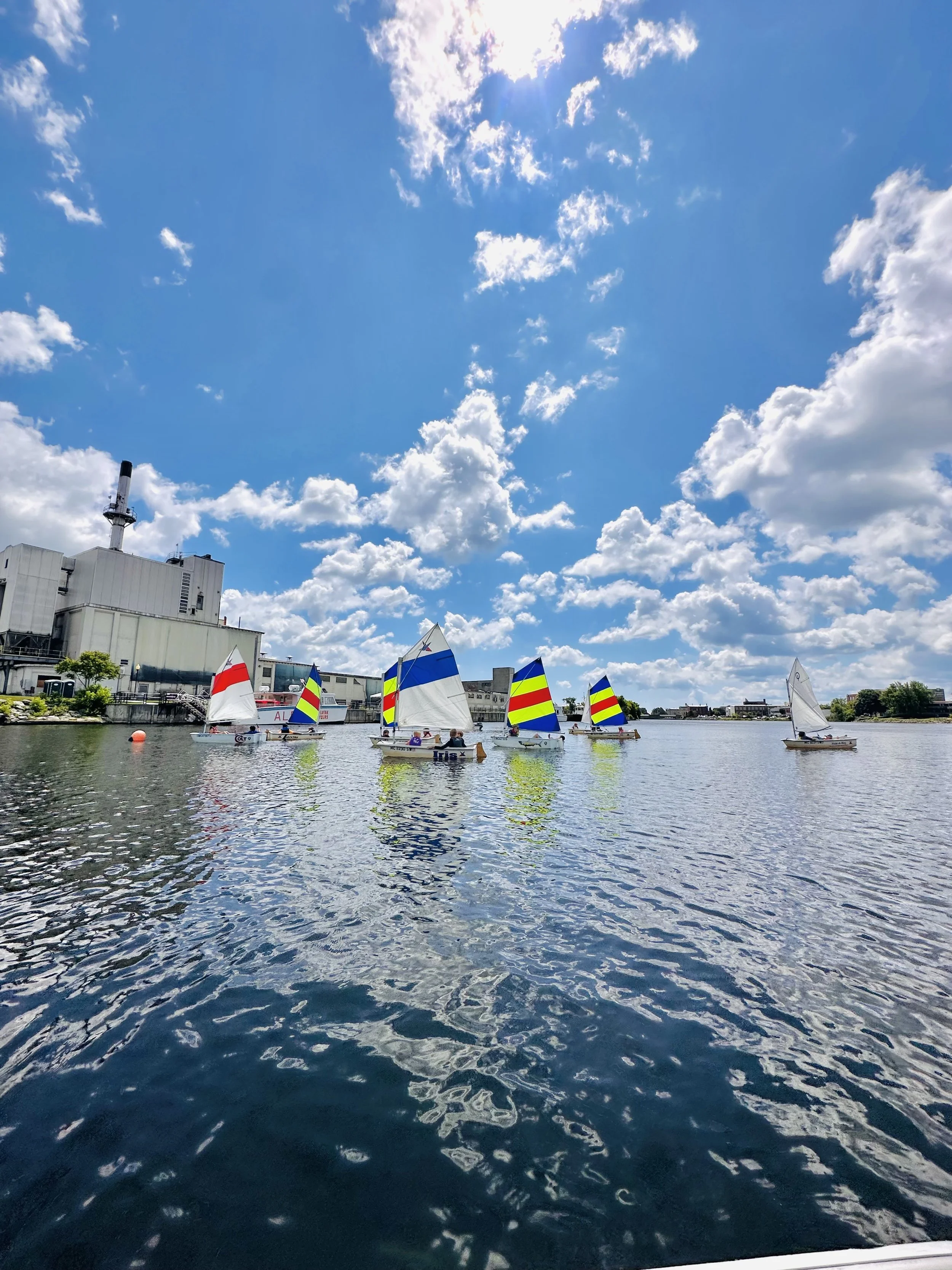 Several small sailboats with colorful striped sails on a calm body of water under a bright blue sky with scattered clouds, near a industrial building.