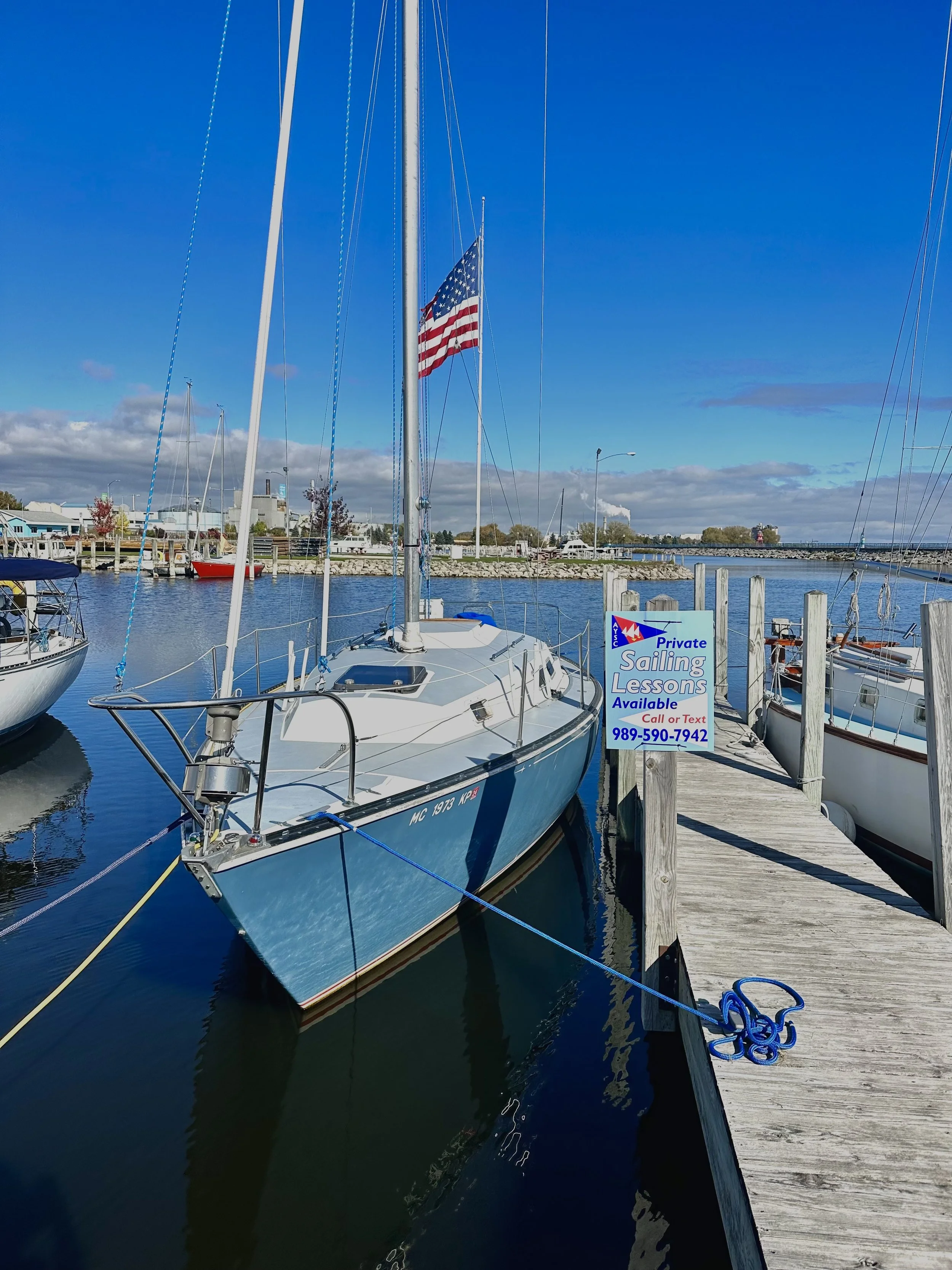 A blue sailboat docked at a marina with an American flag. Sign on dock advertising private sailing lessons with phone number 989-590-7942. Clear blue sky and calm water with other boats in background.
