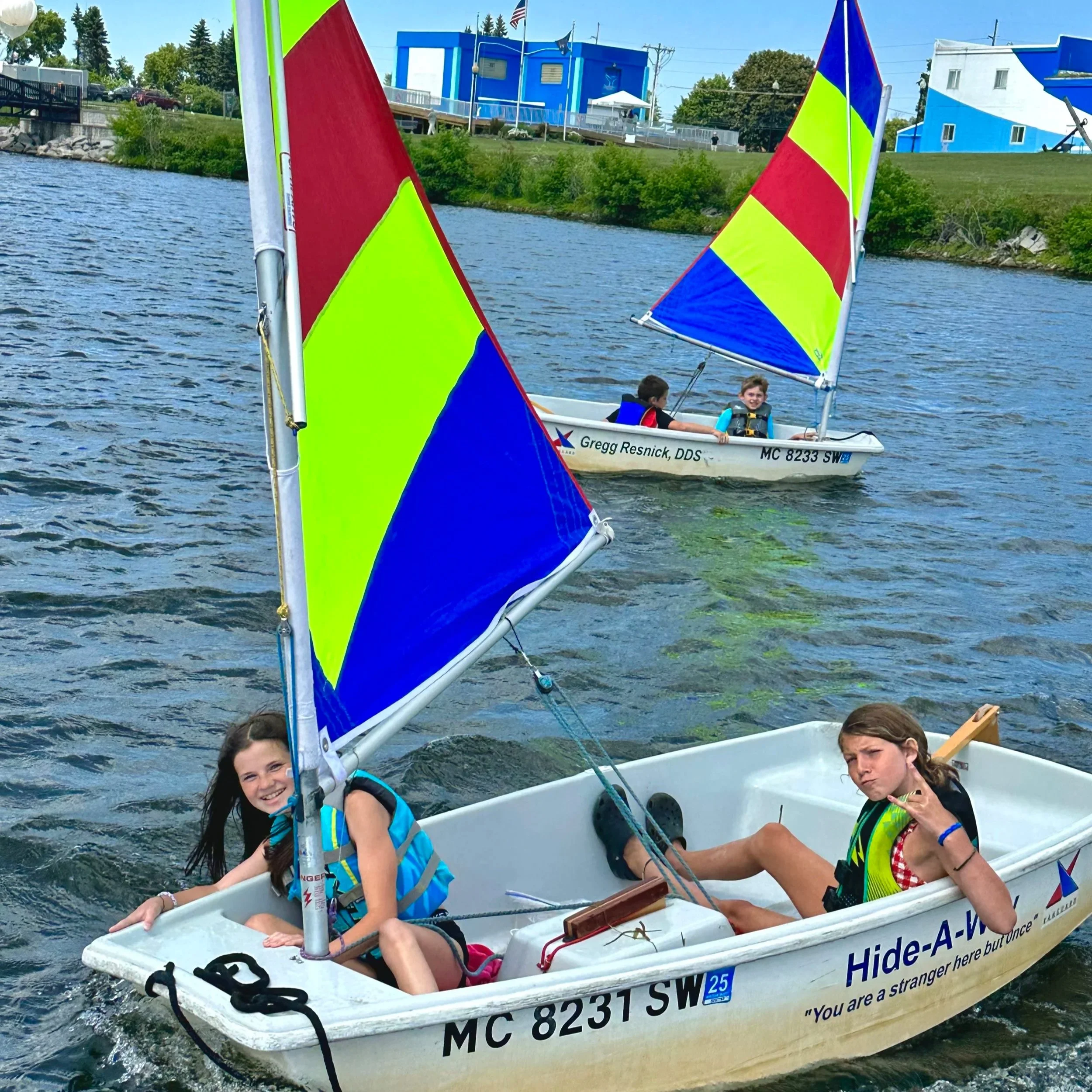 Two children in a small white sailboat, smiling and making peace signs, while another boat with two children sails nearby on a lake. The sailboats have colorful sails and are labeled with registration numbers and the name 'Hide-A-Way' on the boat in the foreground.