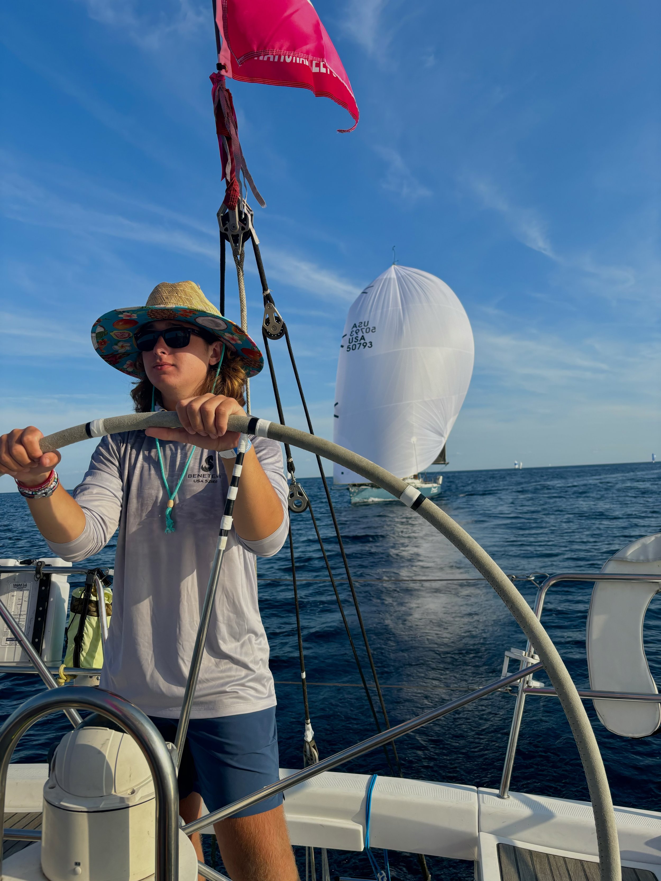 A young person sailing on a boat on the water, wearing a wide-brimmed hat, sunglasses, and a long-sleeve shirt, with a large sailboat with white sails in the background on a clear, sunny day.