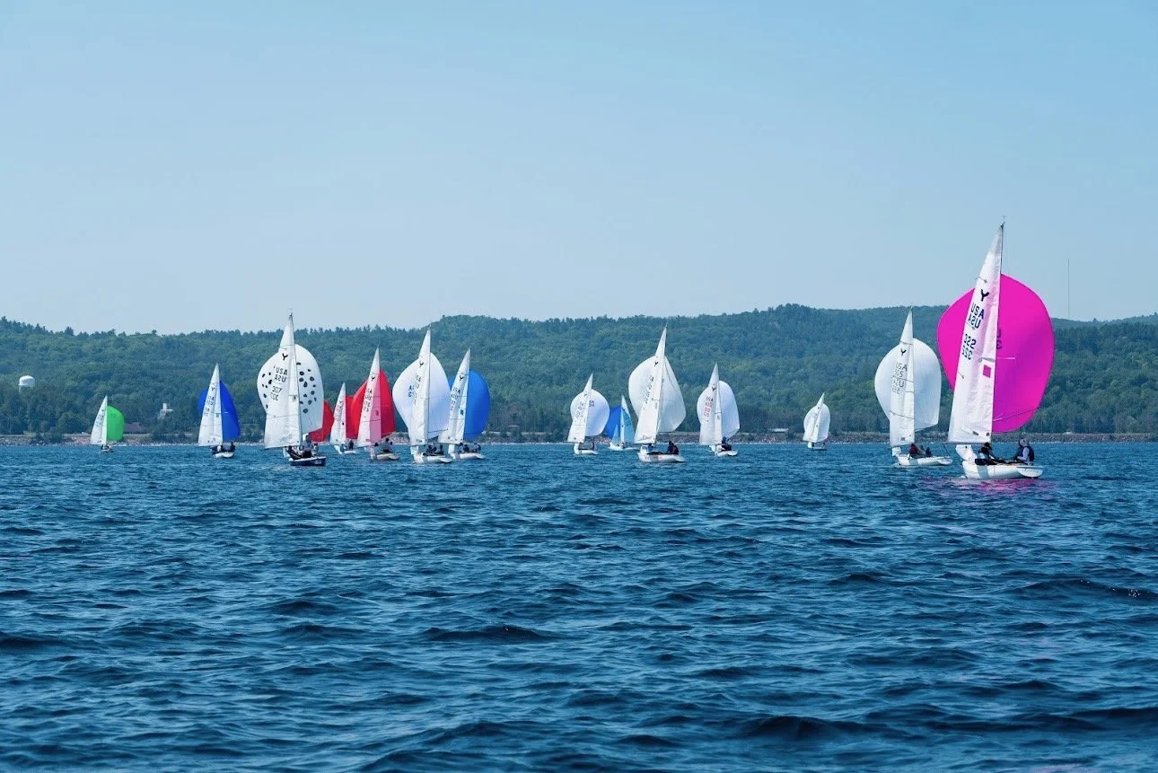 Multiple sailboats with colorful spinnakers racing on a lake with a green, hilly shoreline in the background.