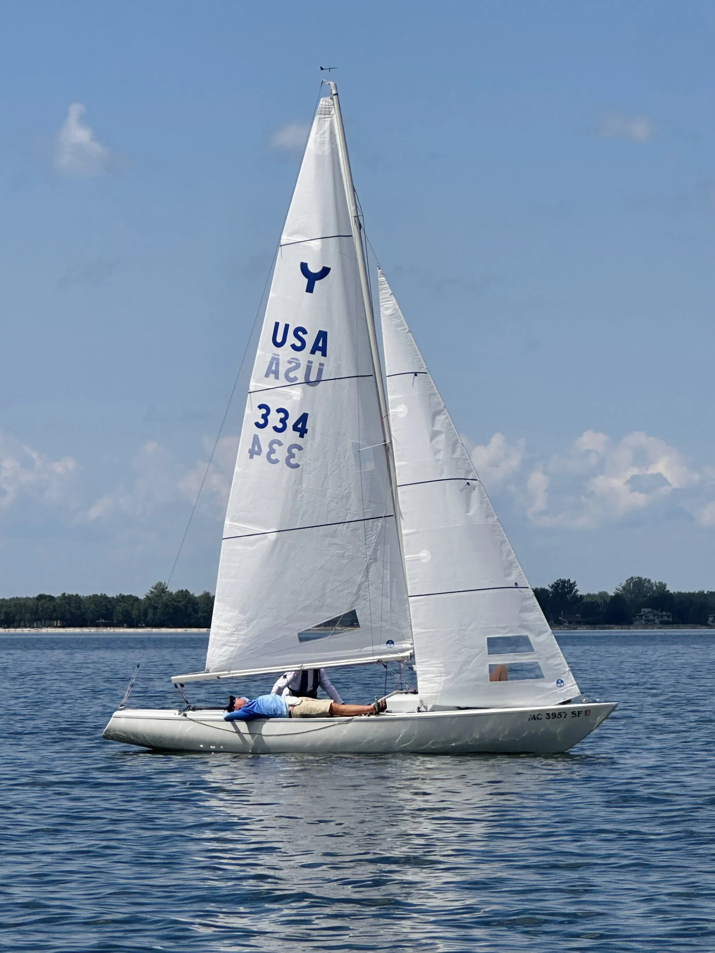 A person lying on a sailboat with sails marked USA and the number 334, on a calm body of water under a partly cloudy sky.