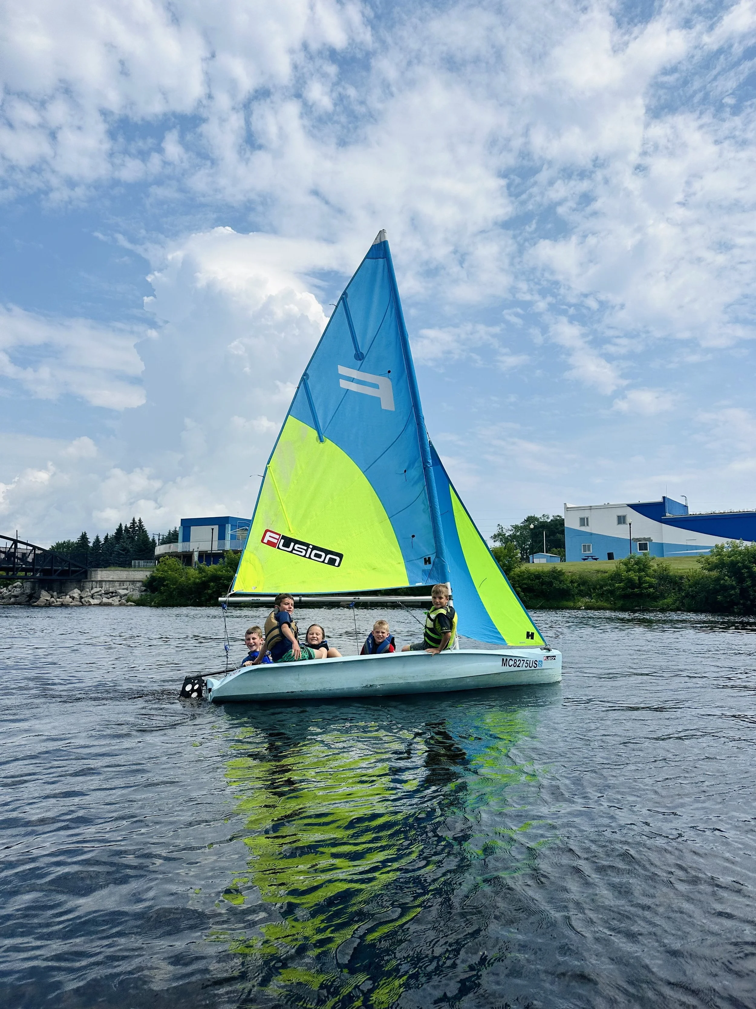 Children sailing on a small boat with a colorful sail on a lake under a partly cloudy sky.