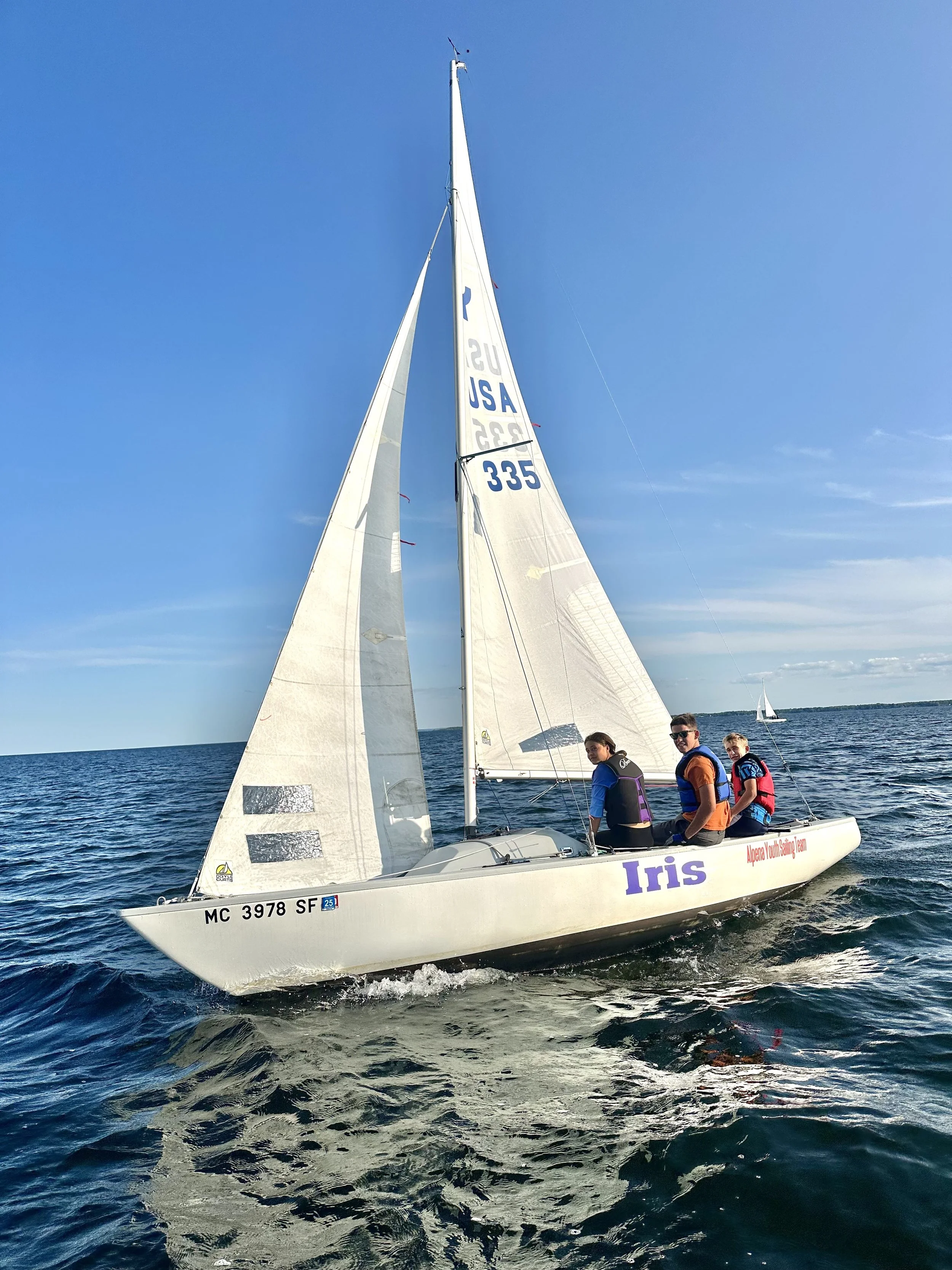 Three people sailing on a white sailboat named Iris on a calm body of water, with one sailboat in the distance, under a clear blue sky.