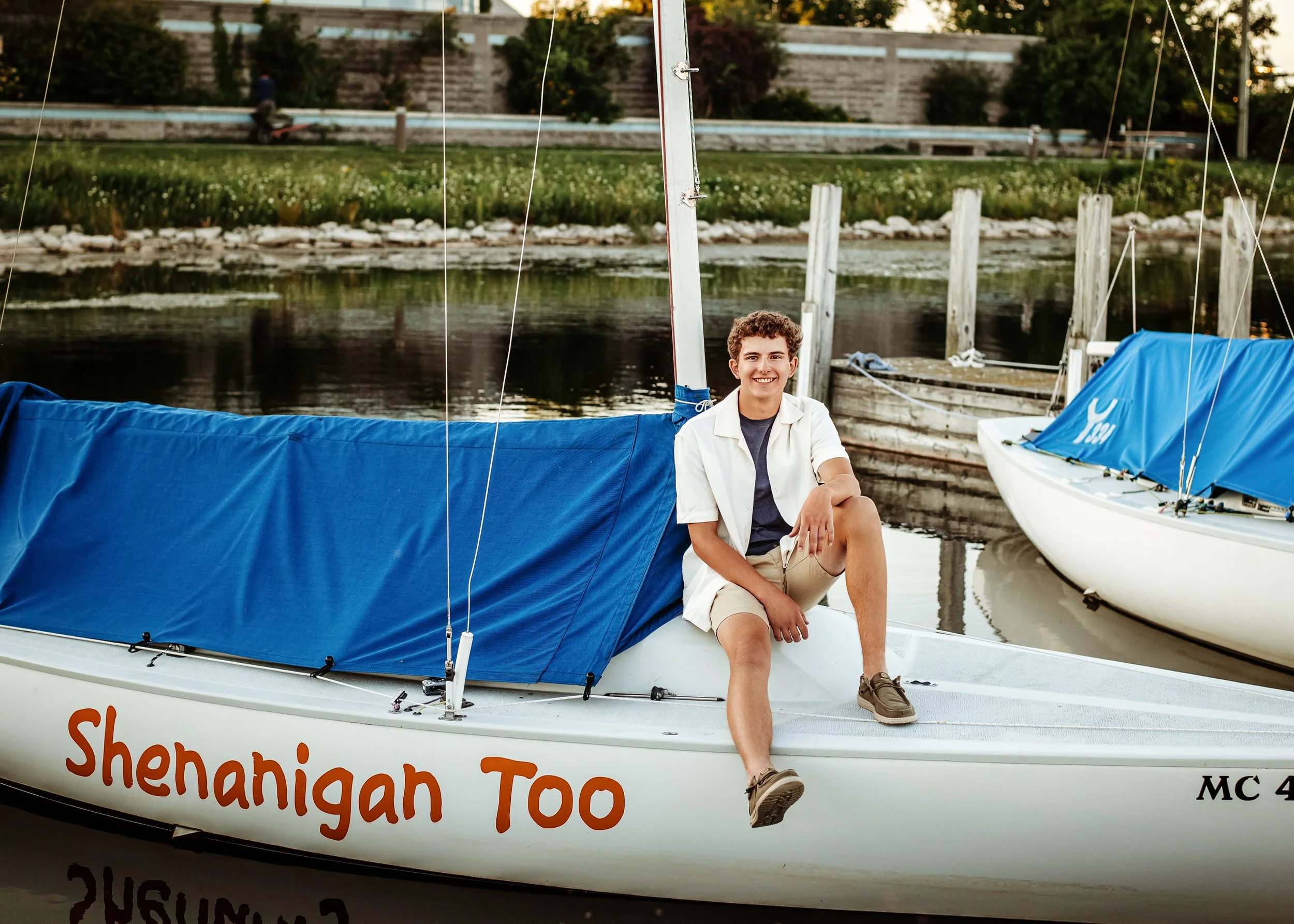A young man sitting on the edge of a sailboat named 'Shenian Too' docked on a calm water body. He is smiling, wearing a white shirt and khaki shorts, with another sailboat visible nearby. In the background, there is a grassy area with trees, a person walking, and a stone wall.