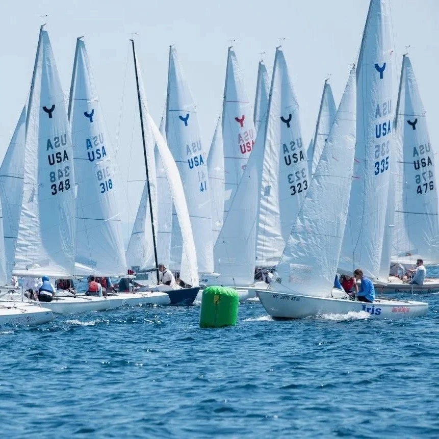Multiple sailboats racing in a regatta on the water, with a green buoy in the foreground.