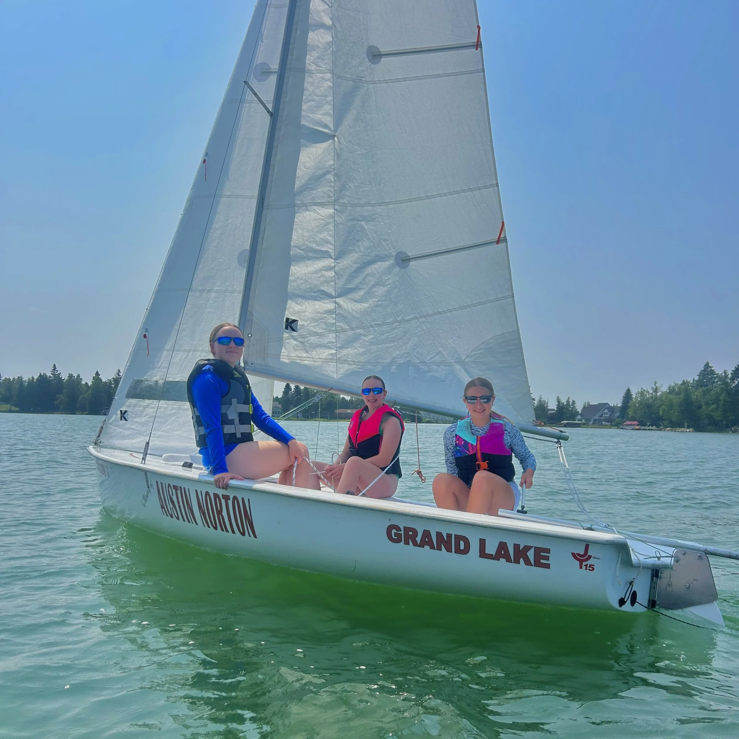 Three women with sunglasses and life jackets sailing on a boat with a large sail on a lake with green water, trees, and houses in the background.