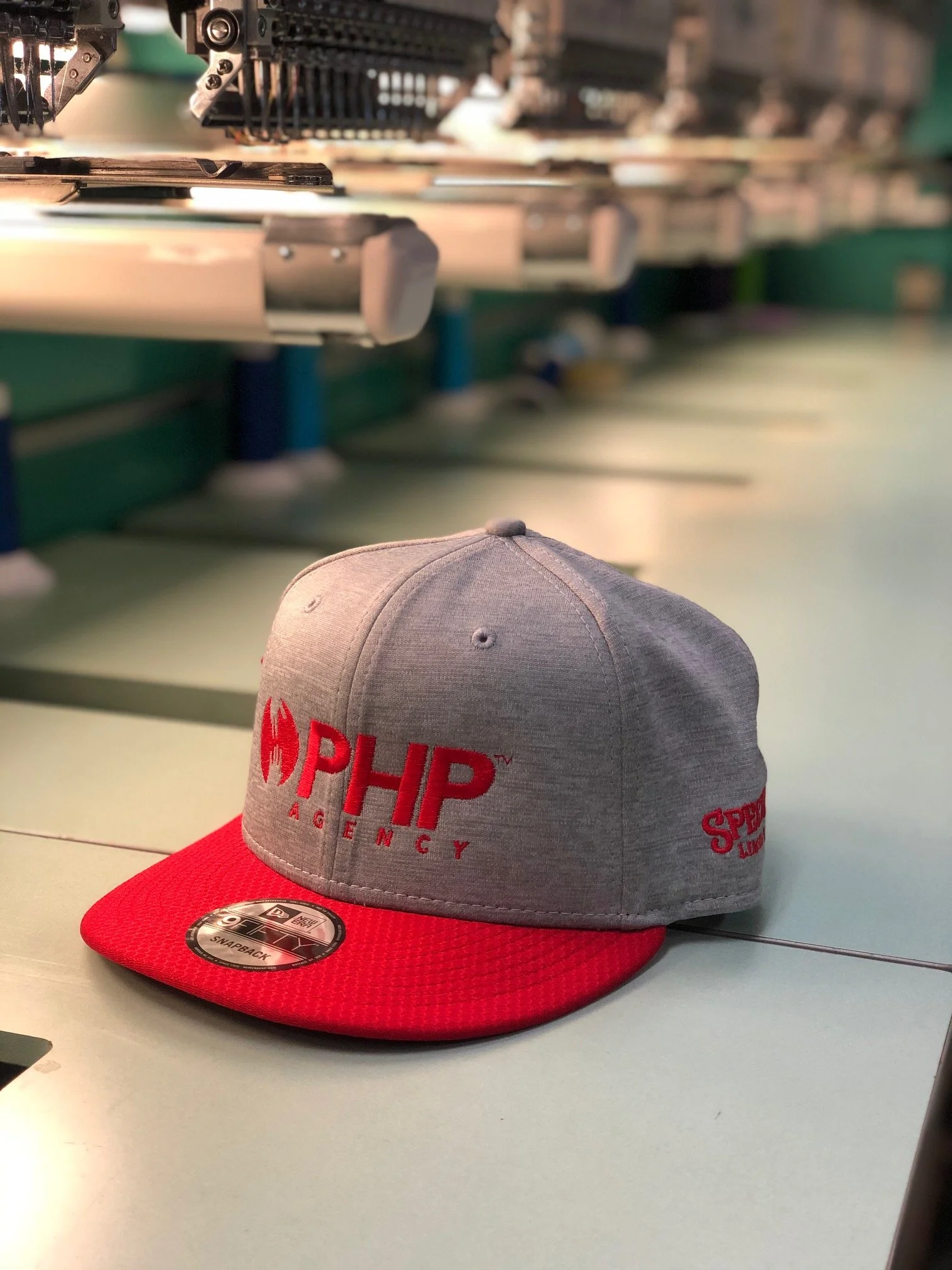 Gray and red baseball cap with "PHP Agency" logo on the front and side, placed on a work table in a local embroidery shop.