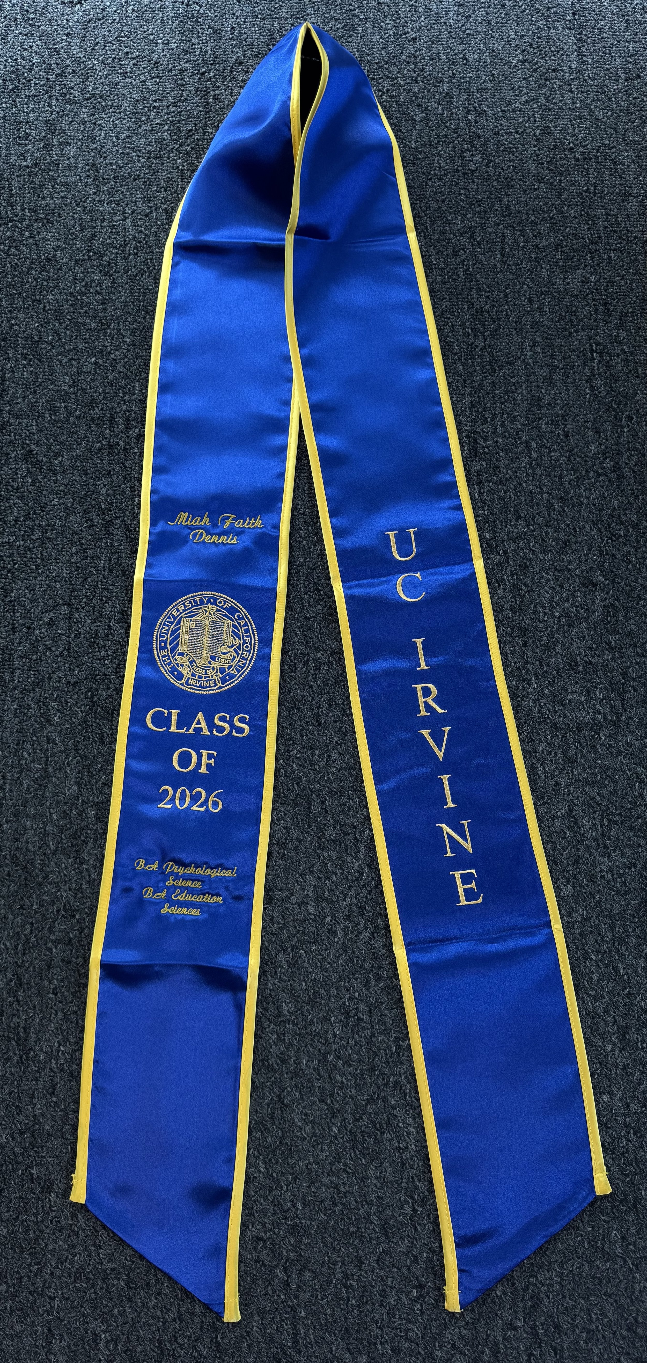 A blue graduation stole with yellow trim. It has personal embroidery that reads "Miah Faith Dennis" on one side and "UC Irvine" on the other. Also includes the UC Irvine seal, the words "Class of 2026," and the phrase "Bet Psychological Science, Bet 