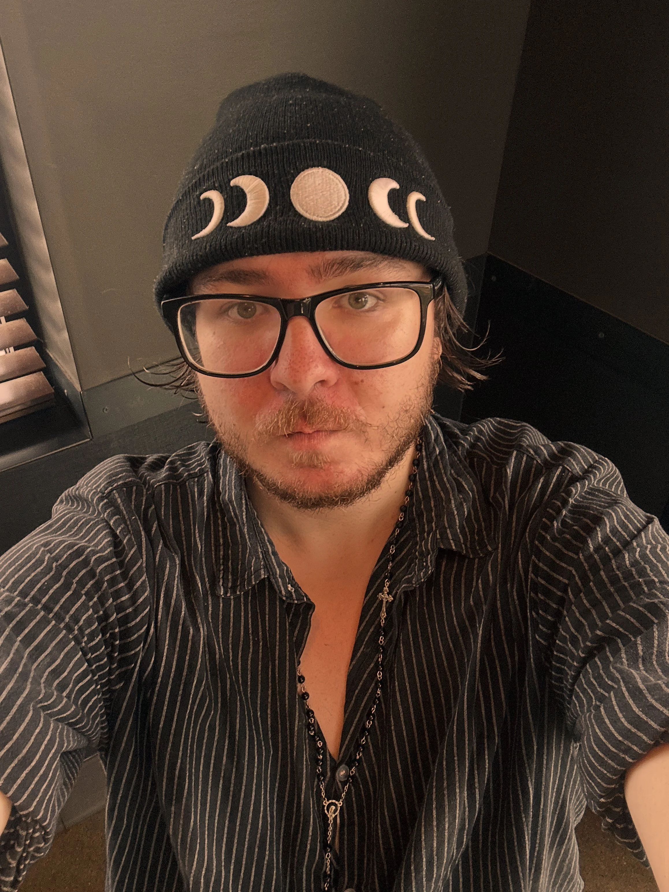 A man with glasses, facial hair, and wearing a black beanie with moon phases, taking a selfie from above. He is wearing a black and gray striped shirt with a beaded necklace featuring a cross.