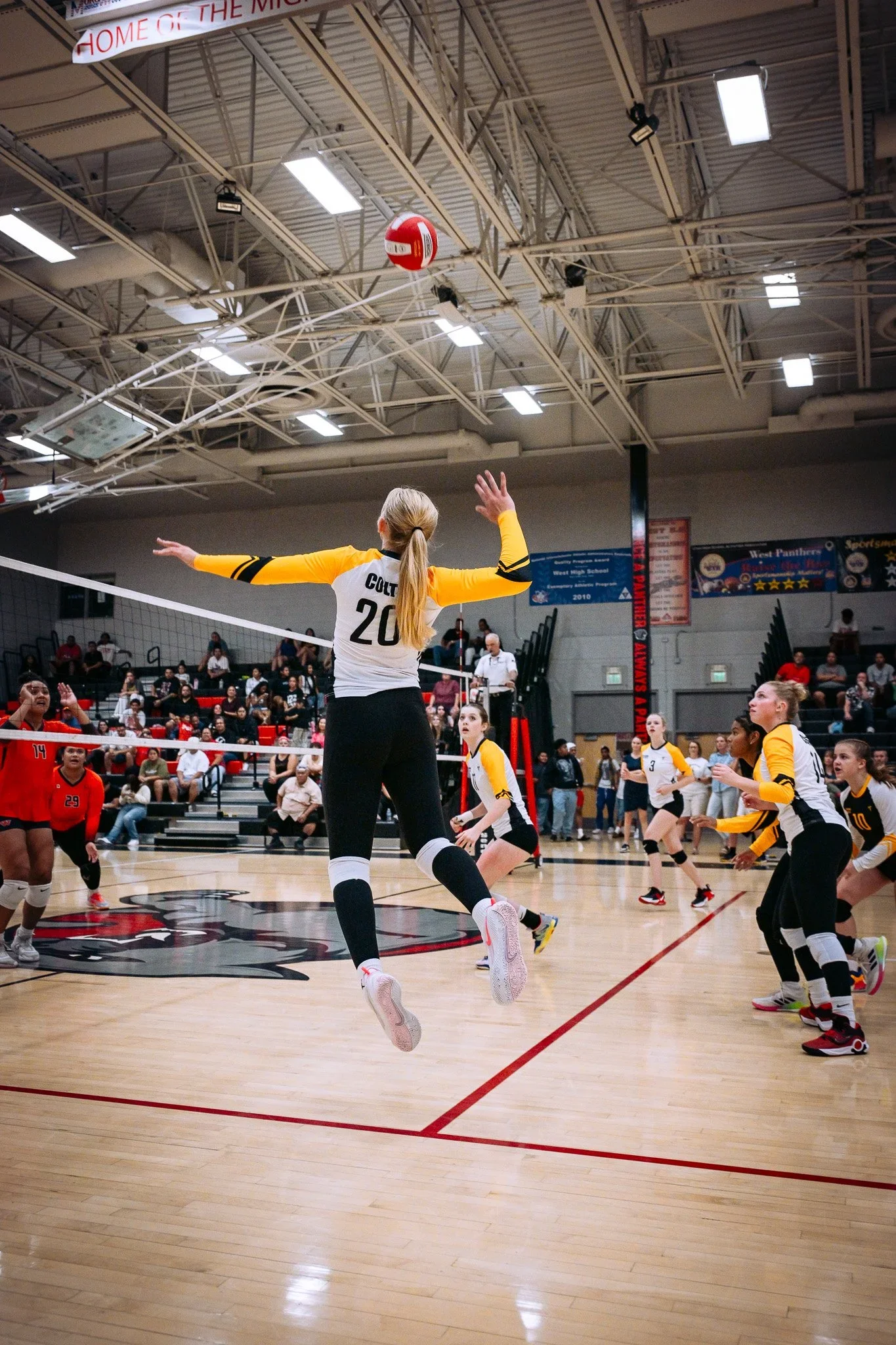 A girls' volleyball game underway in a gymnasium, with players wearing yellow and black and red uniforms. One girl is jumping to block or spike the ball near the net, with spectators watching from the bleachers in the background.
