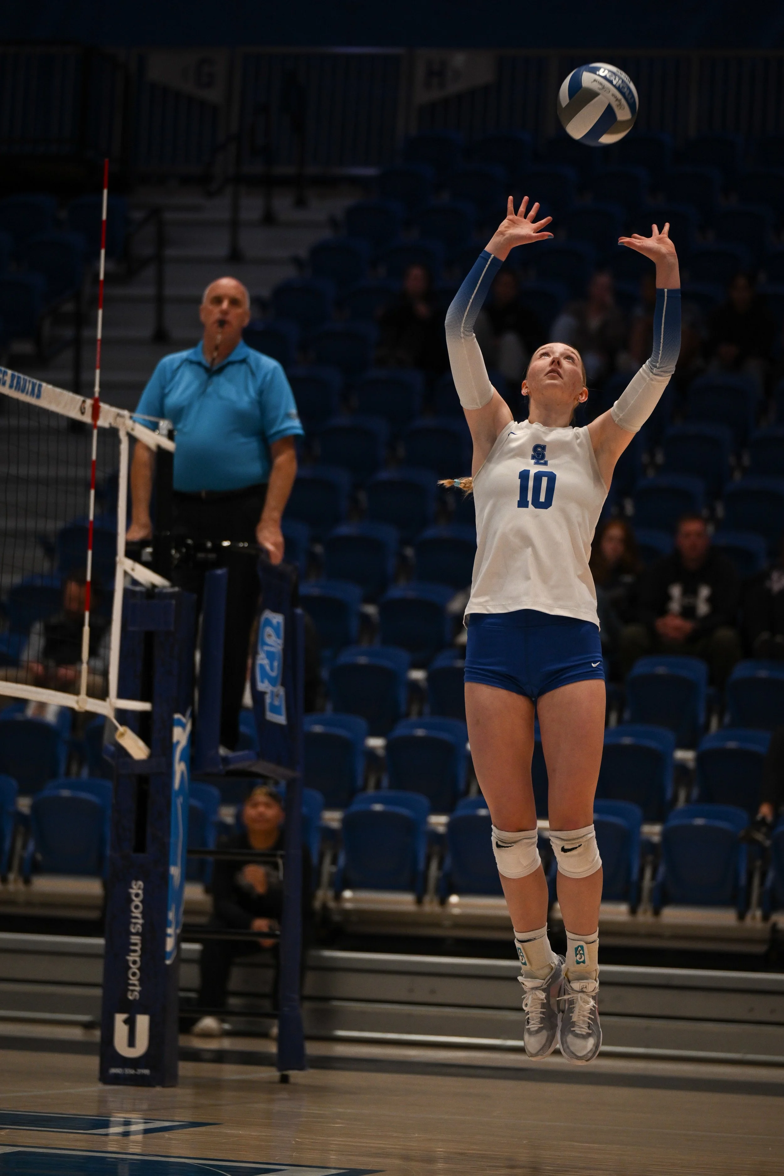 A female volleyball player with jersey number 10 is jumping to set the ball at an indoor volleyball court. An official in a blue shirt is watching nearby, with empty blue seats in the background.