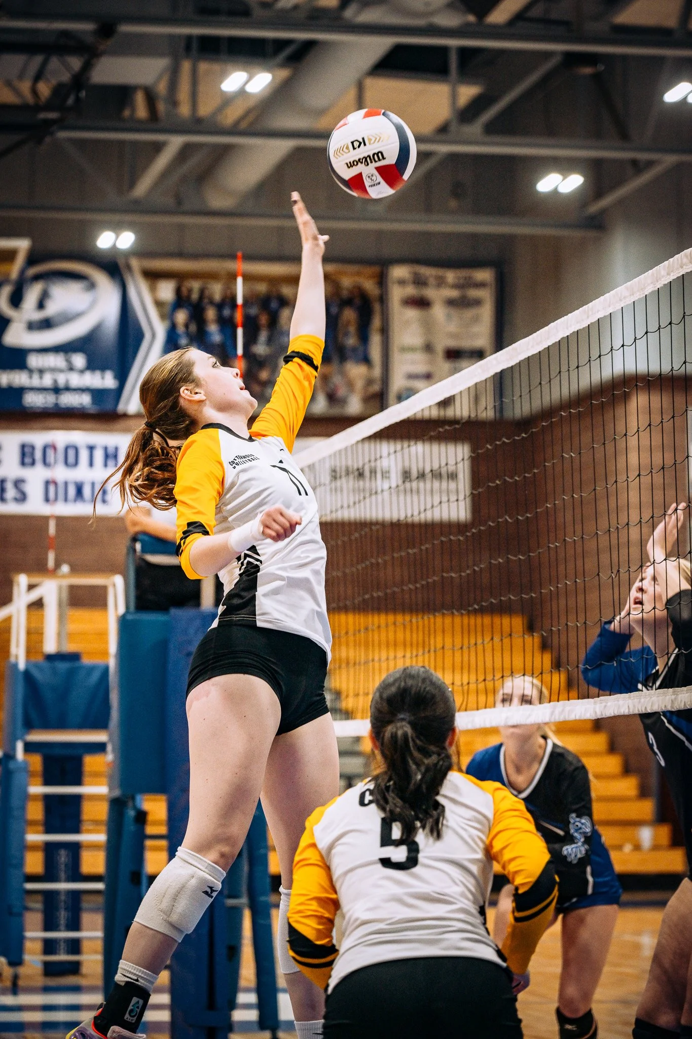 A female volleyball player in a white and yellow jersey jumping to hit a volleyball over the net during a match in an indoor gymnasium.