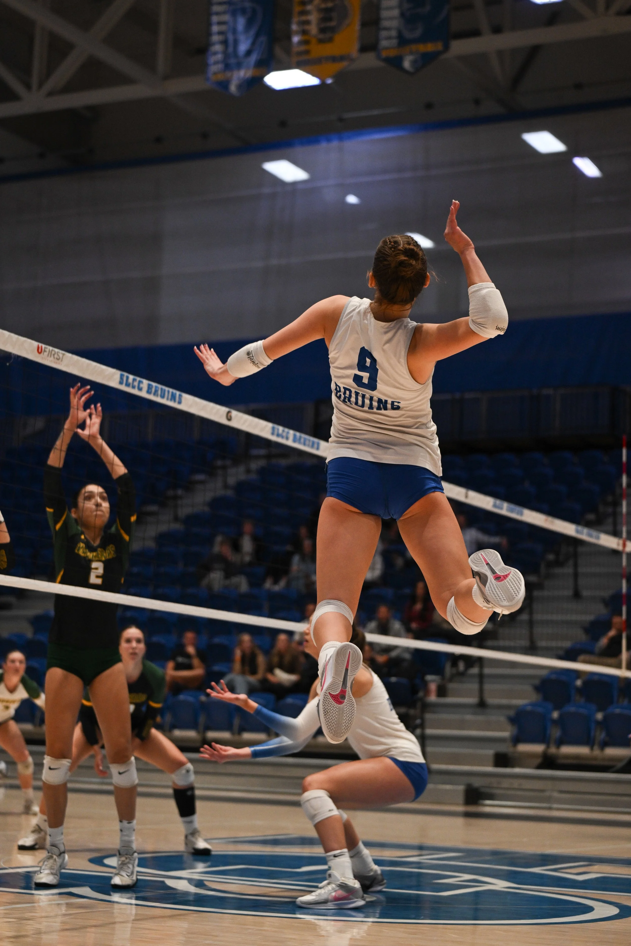 A female volleyball player with jersey number 9 from the SLEC Bruins team jumping to spike the ball over the net during a volleyball match, with her teammates and opponents on the court.
