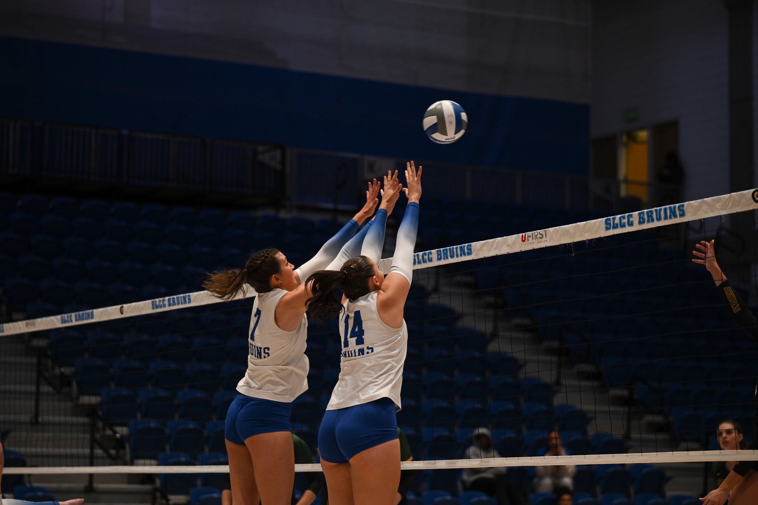 Two volleyball players jumping to block a ball at the net during a game, with empty blue seats in the background.