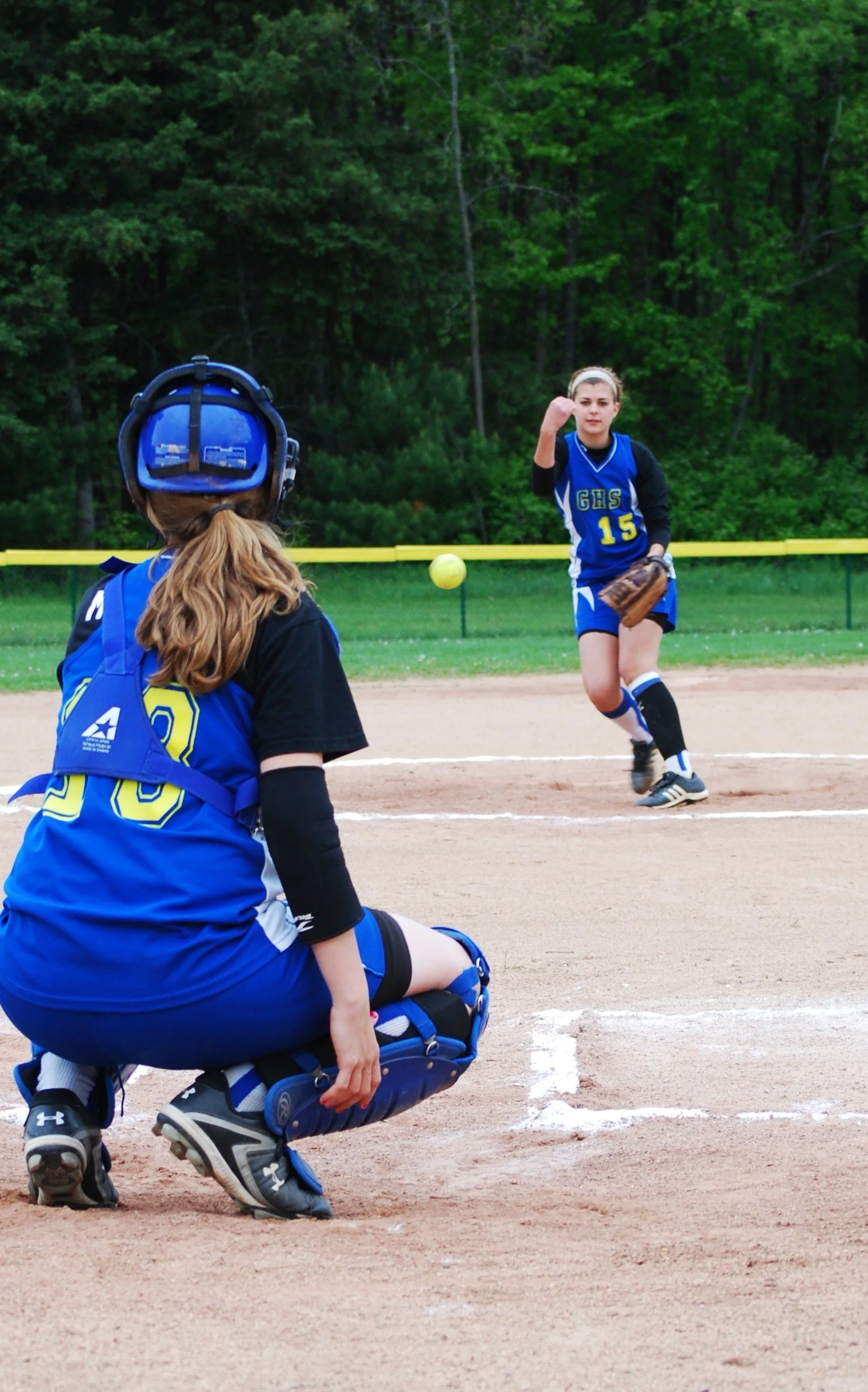 A softball game with one player pitching and another crouched behind home plate wearing catcher gear, on a field with green trees in the background.