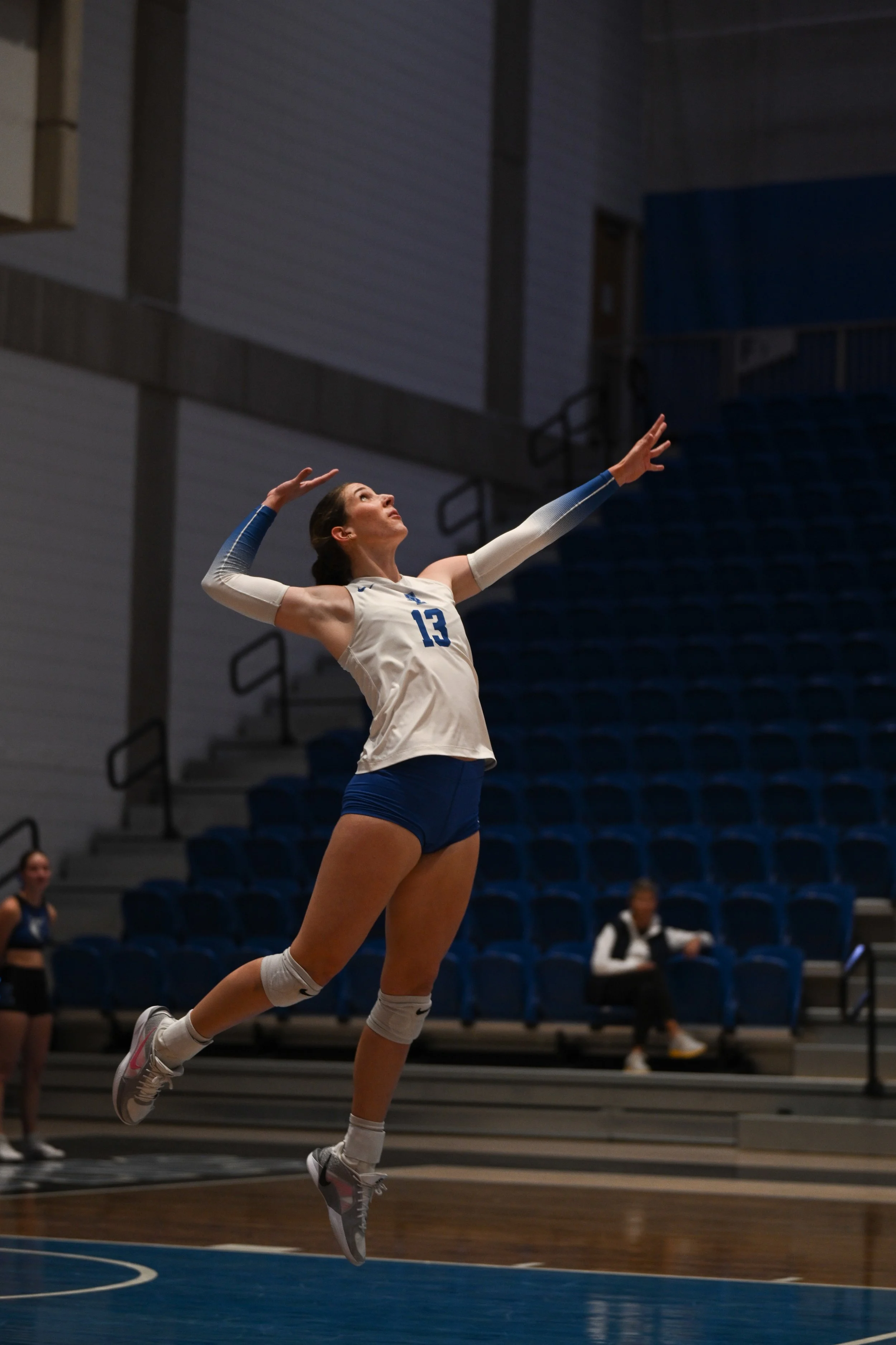 A female volleyball player in a white jersey with the number 13 is jumping in the air on an indoor court with blue seating in the background.