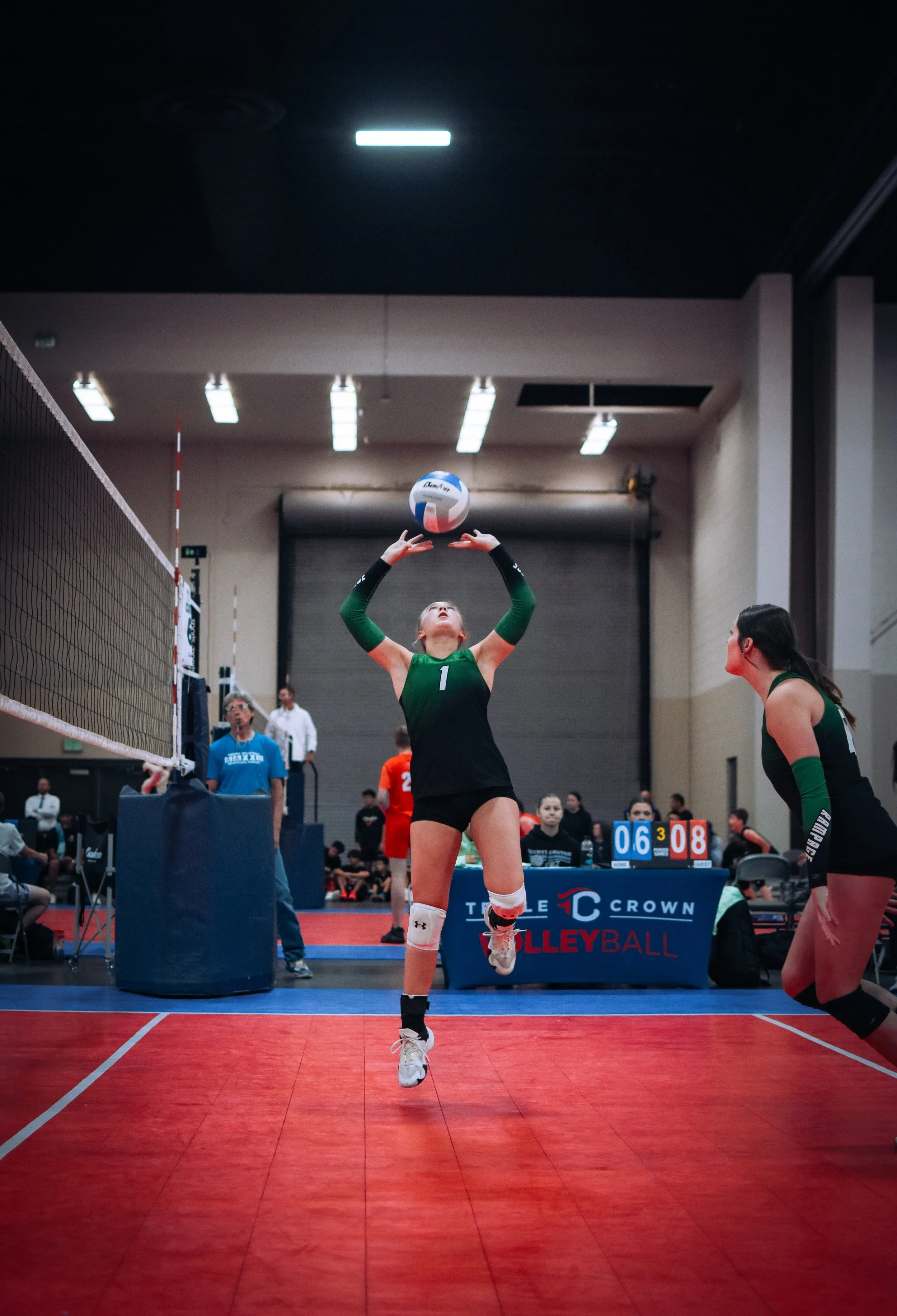 A female volleyball player in a green and black uniform jumping to set the ball during a match in an indoor gym. Another player in green is crouched nearby, and a volleyball net is visible on the left side. The background shows a scoreboard and spect