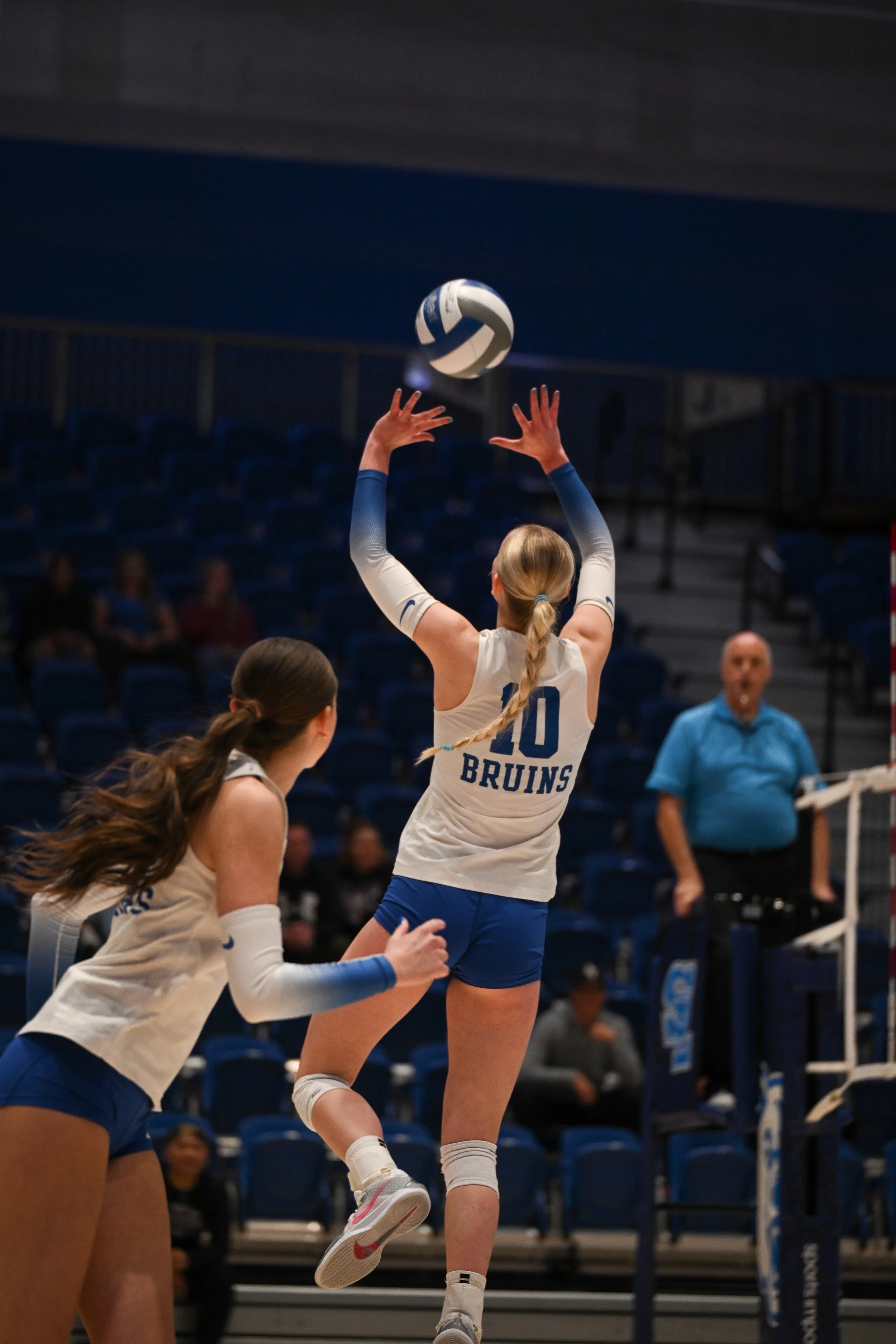 Two female volleyball players in blue and white uniforms are playing on an indoor court. One player is jumping to hit the volleyball, while the other is standing nearby, watching. There are empty bleacher seats and a referee in a blue shirt in the ba