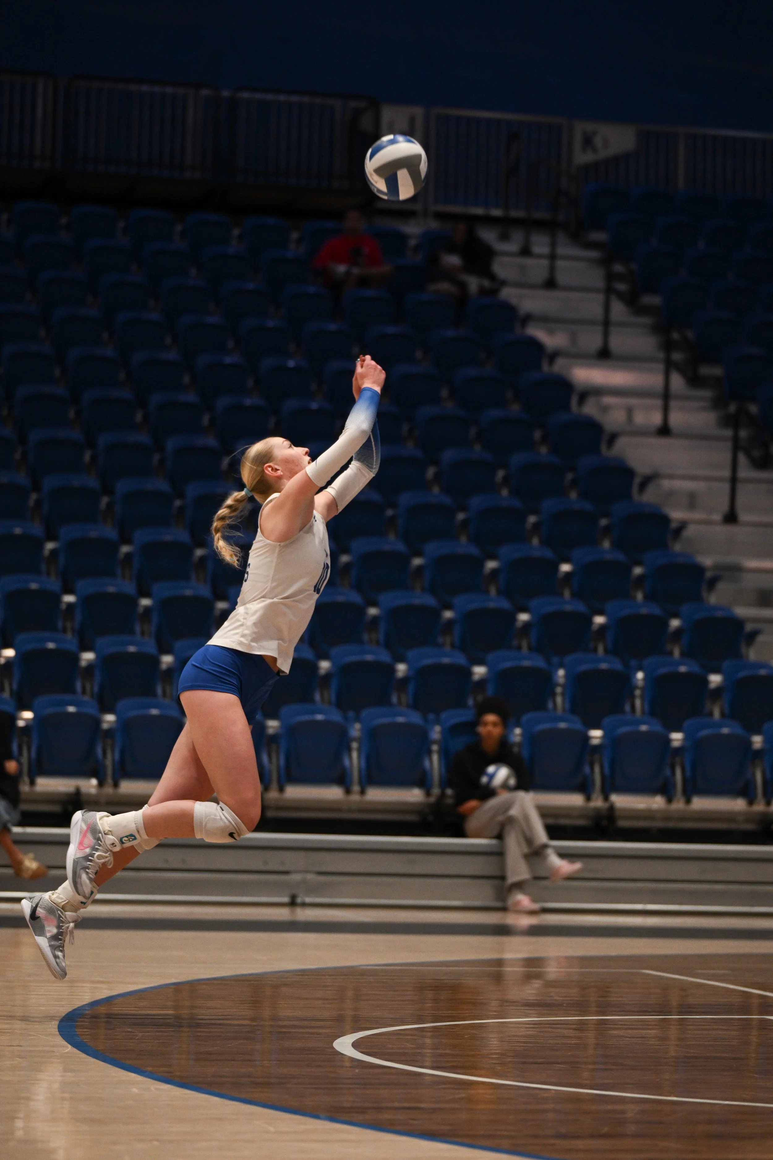 A female volleyball player jumps to hit a volleyball in an indoor gym with blue seats and a few spectators in the background.