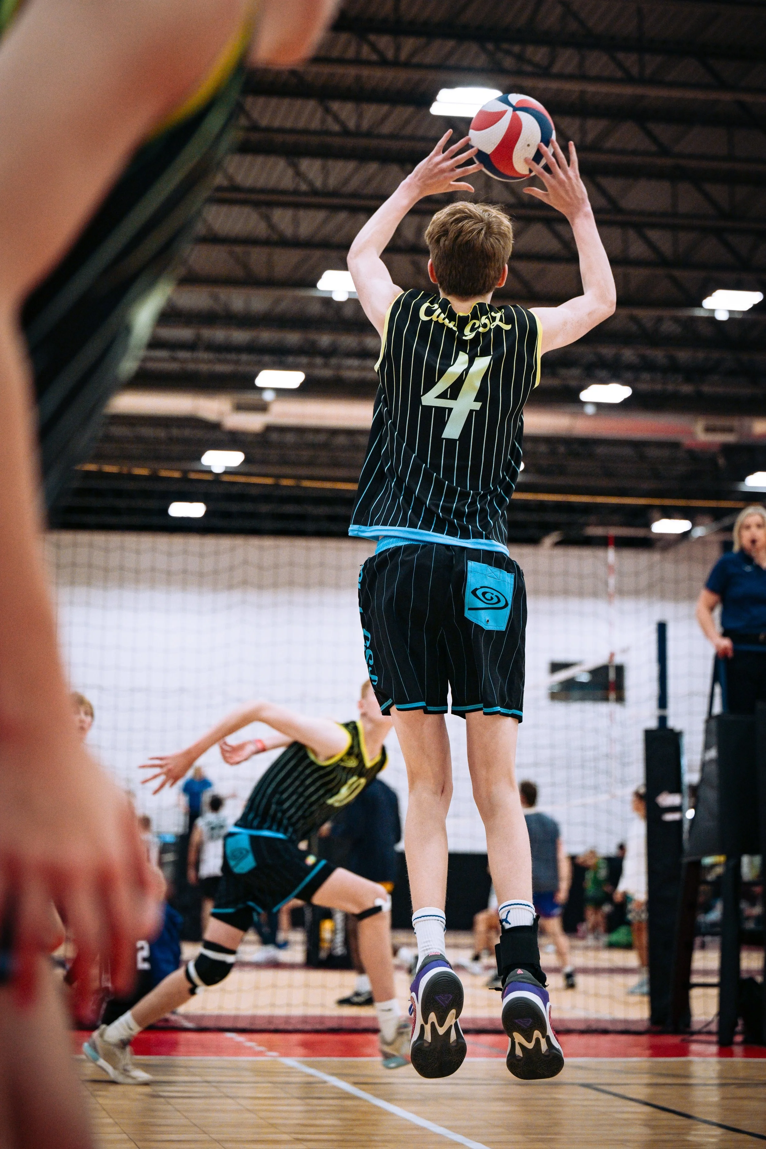 A young male volleyball player wearing a black and blue uniform with the number 4 jumps to set a volleyball during an indoor match.