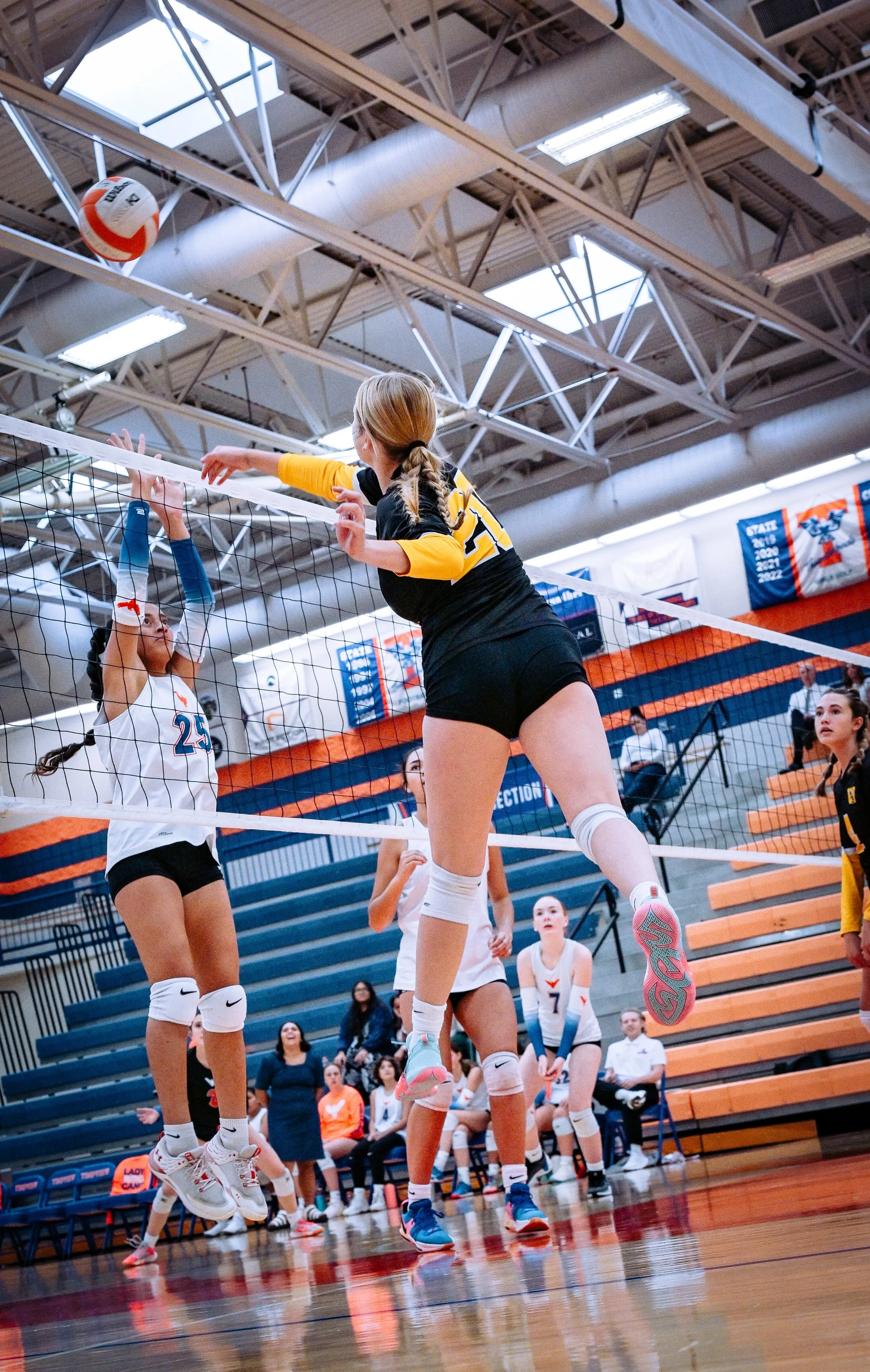 Young female volleyball player in a black and yellow uniform jumping to spike the ball over the net while teammate in white and blue attempts to block.